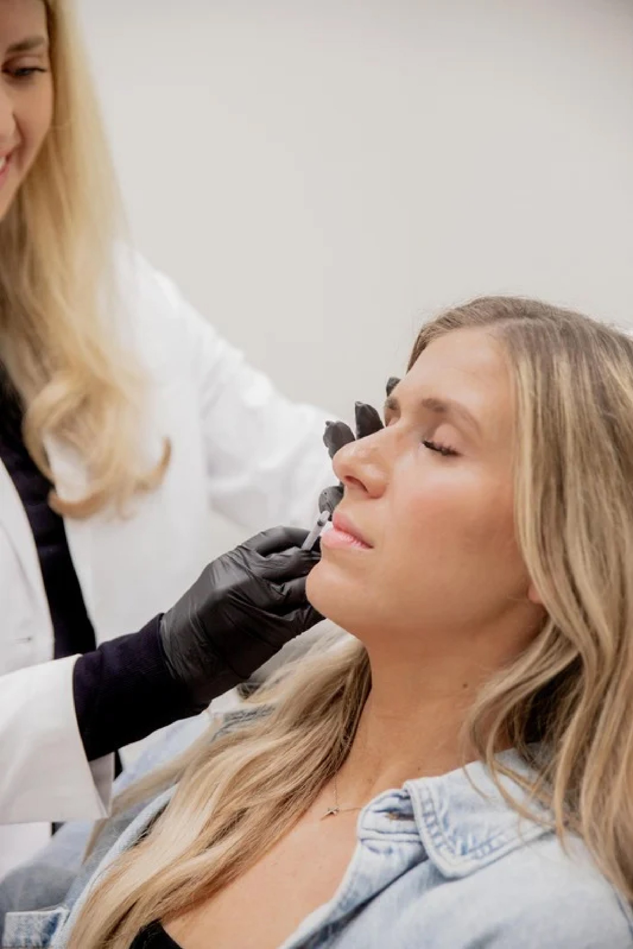 A woman with blonde hair receiving a cosmetic injection from a doctor wearing black gloves in a clinical setting.