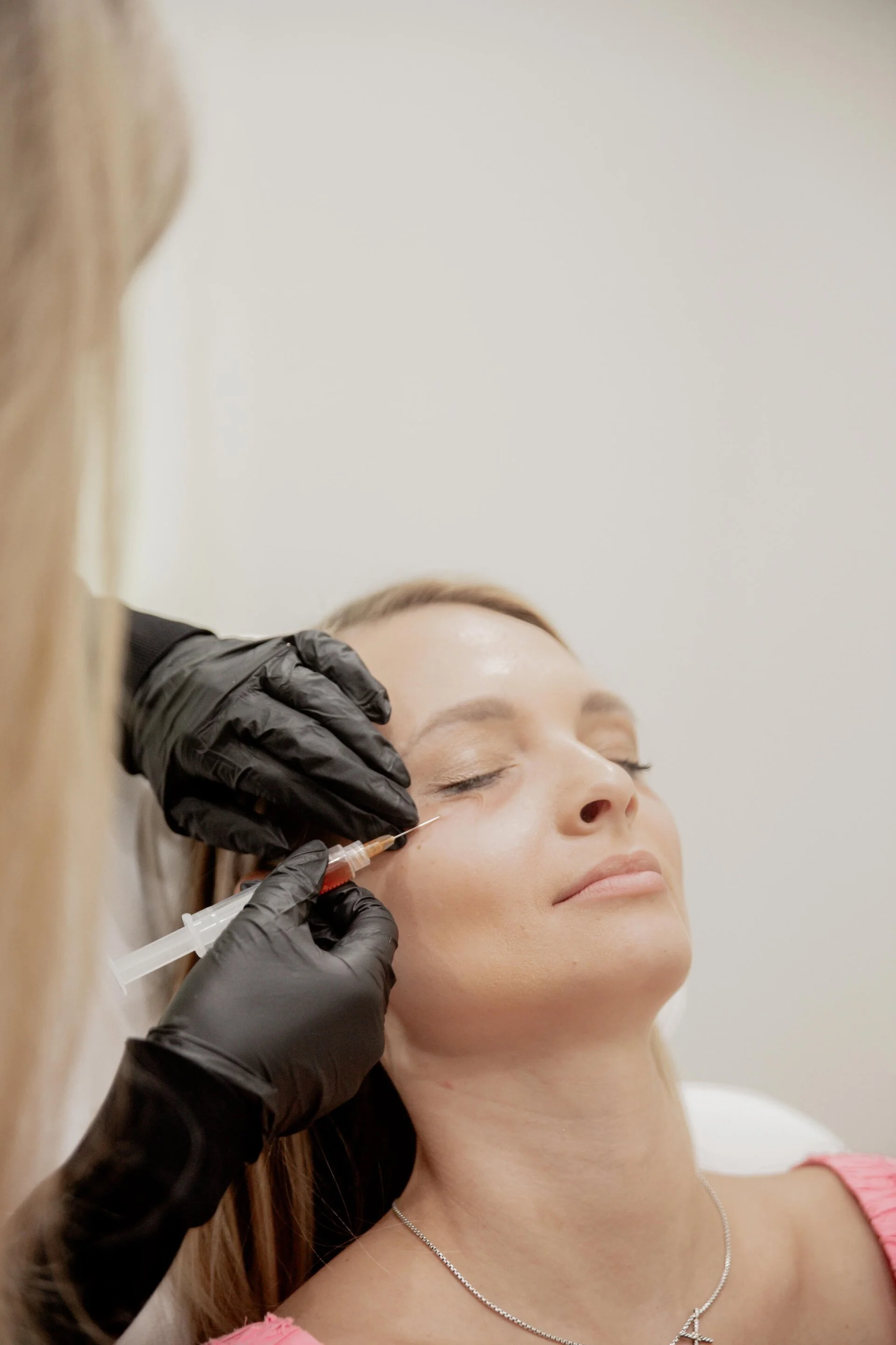A woman receiving a cosmetic injection in her cheek from a medical professional wearing black gloves in a clinical setting.