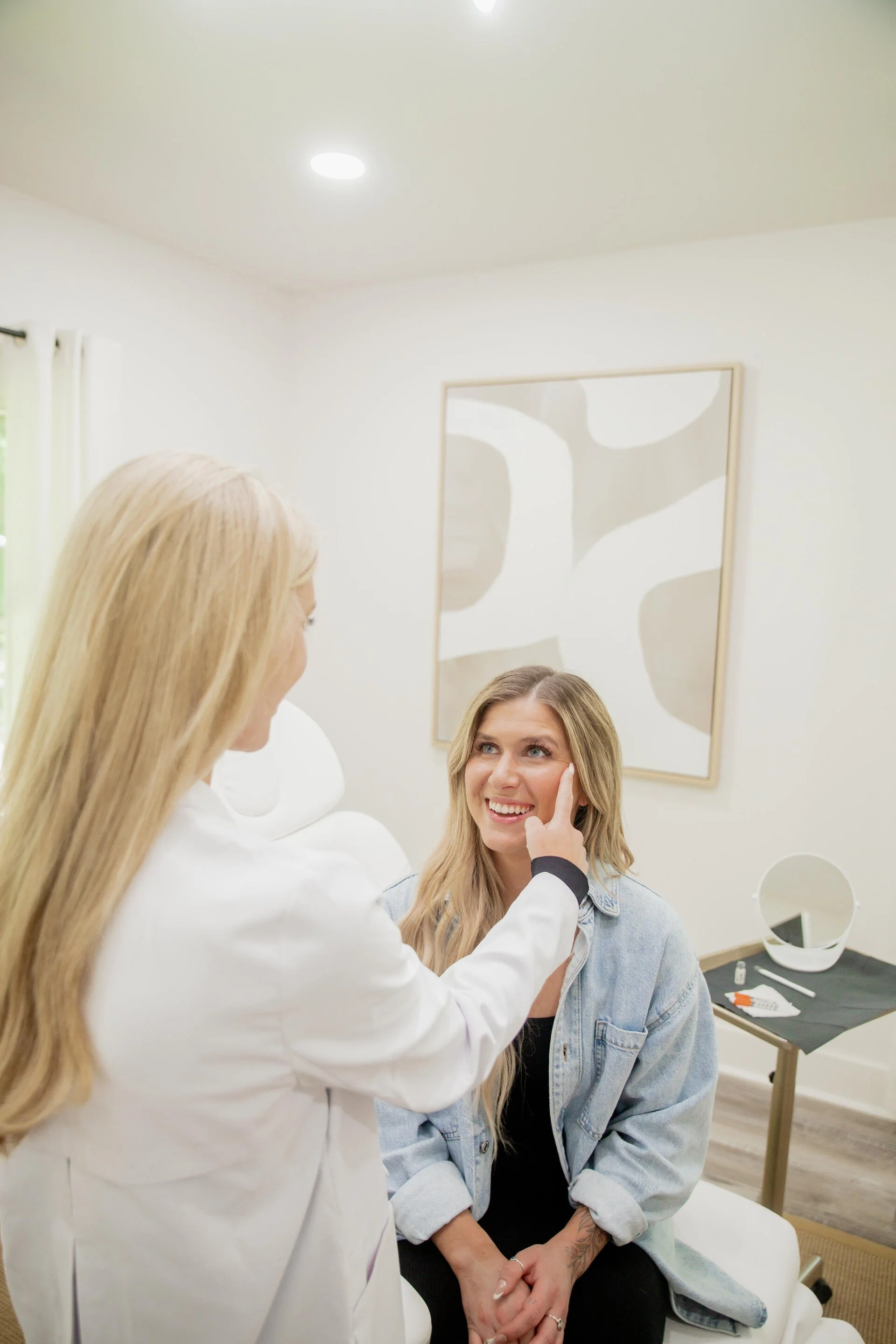 A woman sitting on a medical examination bed in a doctor’s office, smiling as a healthcare professional gently touches her face.