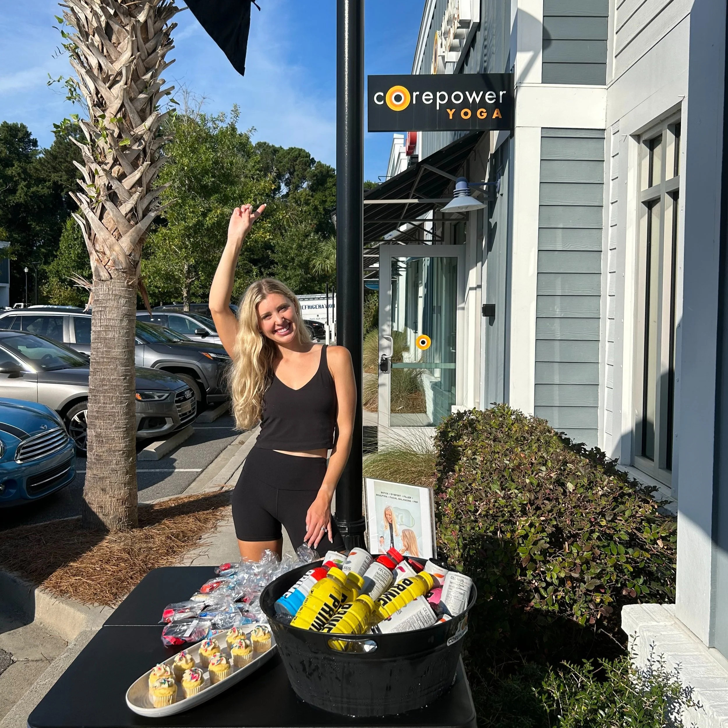 A smiling woman with long blonde hair in a black tank top and shorts stands outdoors next to a table with snacks and bottles, pointing upward.