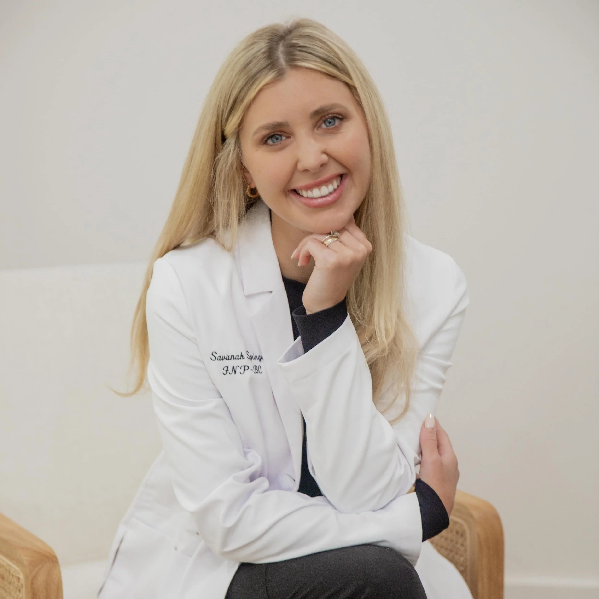 A smiling blonde woman in a white medical coat sitting in an examination room.