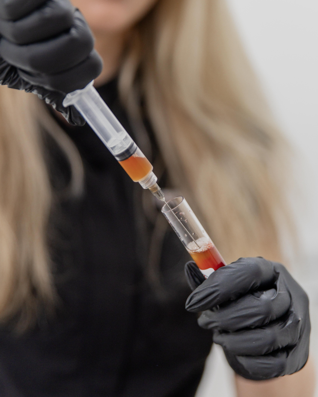 Person wearing black gloves using a syringe to transfer blood sample into a test tube.