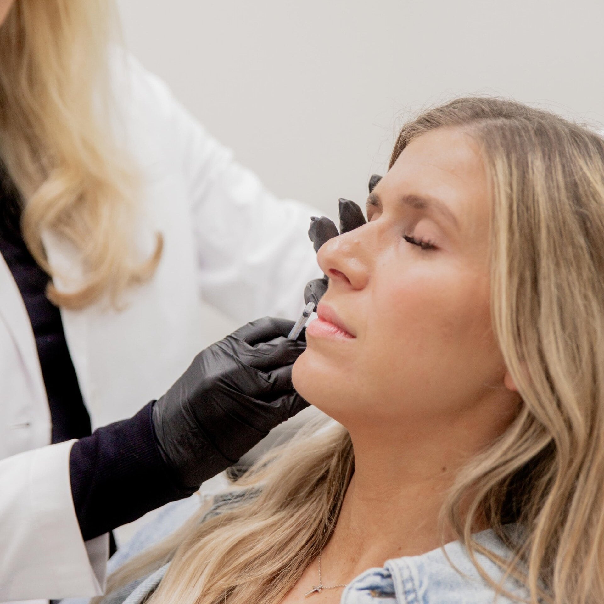 A woman receiving a cosmetic injection to her lips from a healthcare professional wearing black gloves.