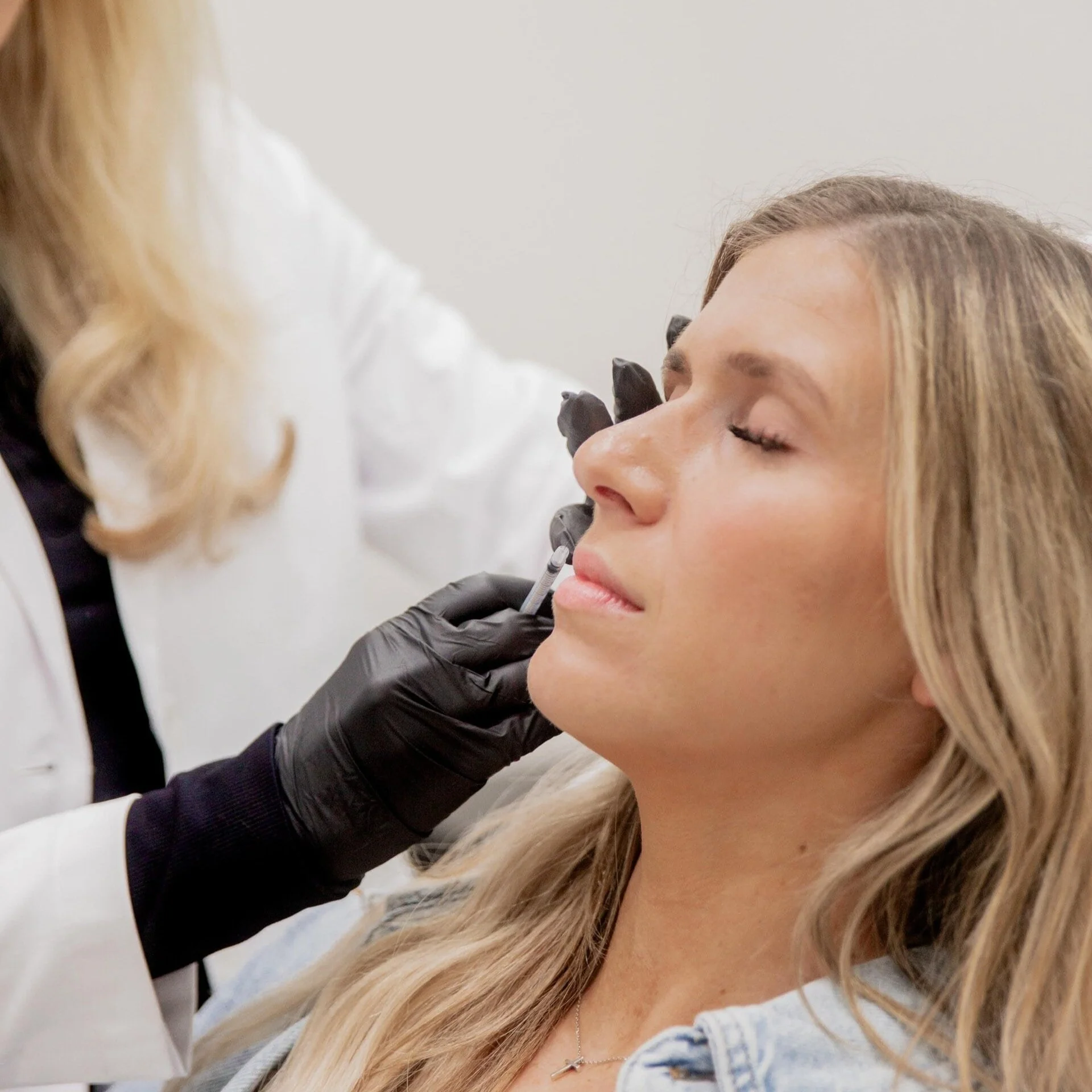 A woman receiving a cosmetic injection on her cheek from a healthcare professional wearing black gloves.