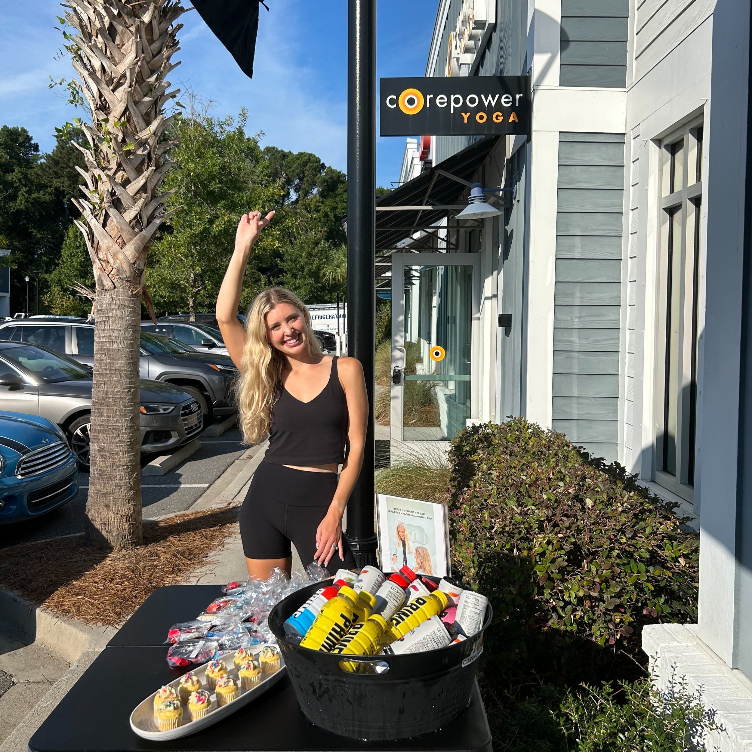 A smiling woman with long blonde hair in a black tank top and shorts standing outside a building with a sign that reads 'corepower YOGA.' She is pointing upwards with her right hand. In front of her is a table with cupcakes and a large container fill