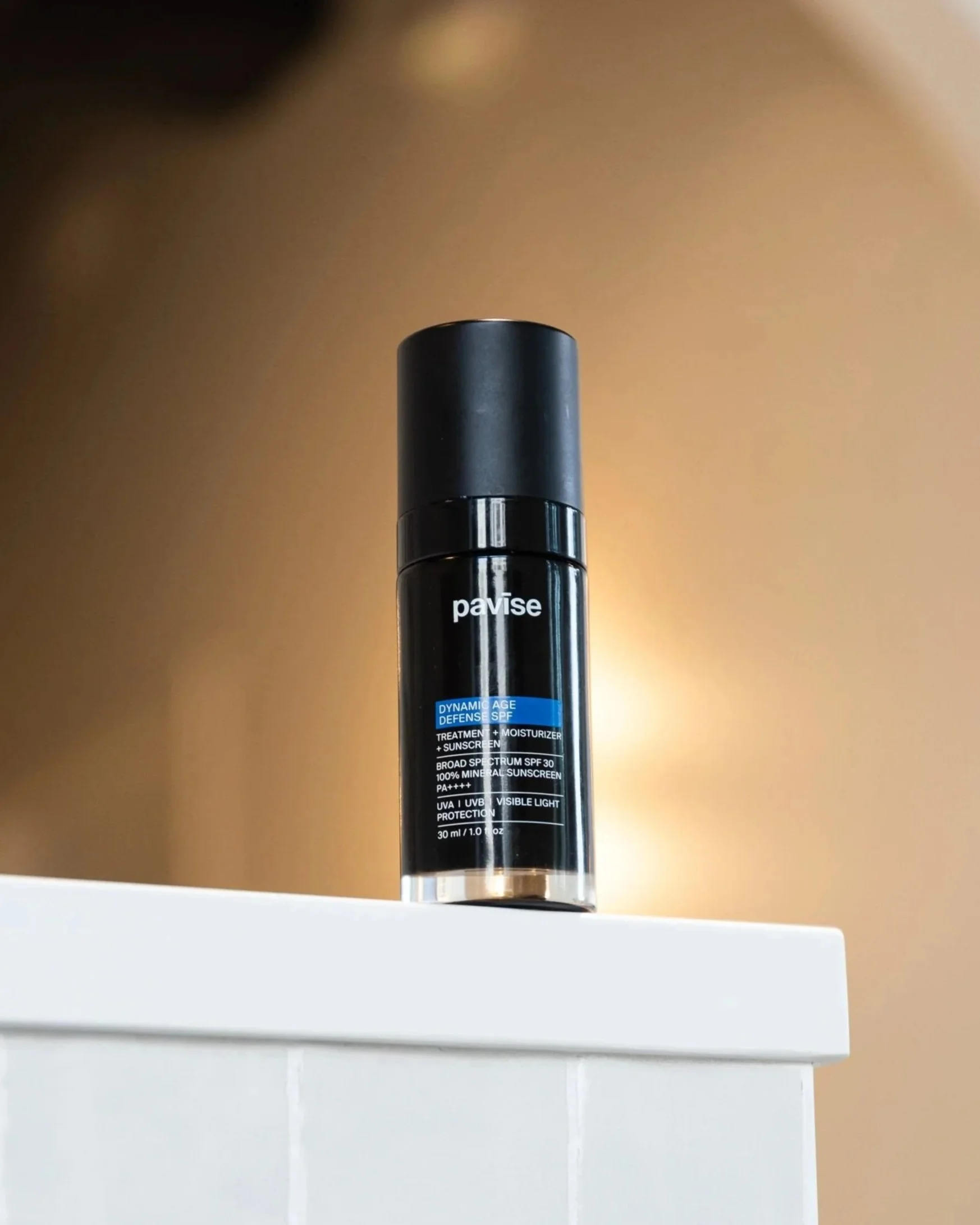 A black bottle of Pavise Dynamic Age Defense SPF sunscreen and moisturizer placed on the white edge of a surface, with a blurred beige background.