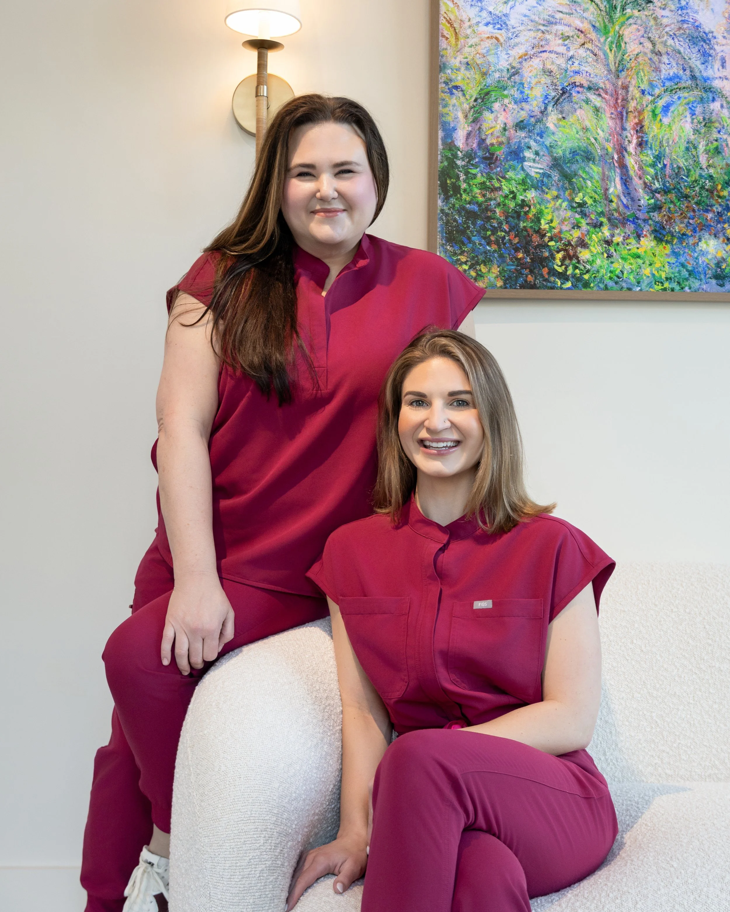 Two women in matching scrubs, one standing and one sitting, smiling in a modern room with a colorful abstract painting and a wall-mounted lamp.
