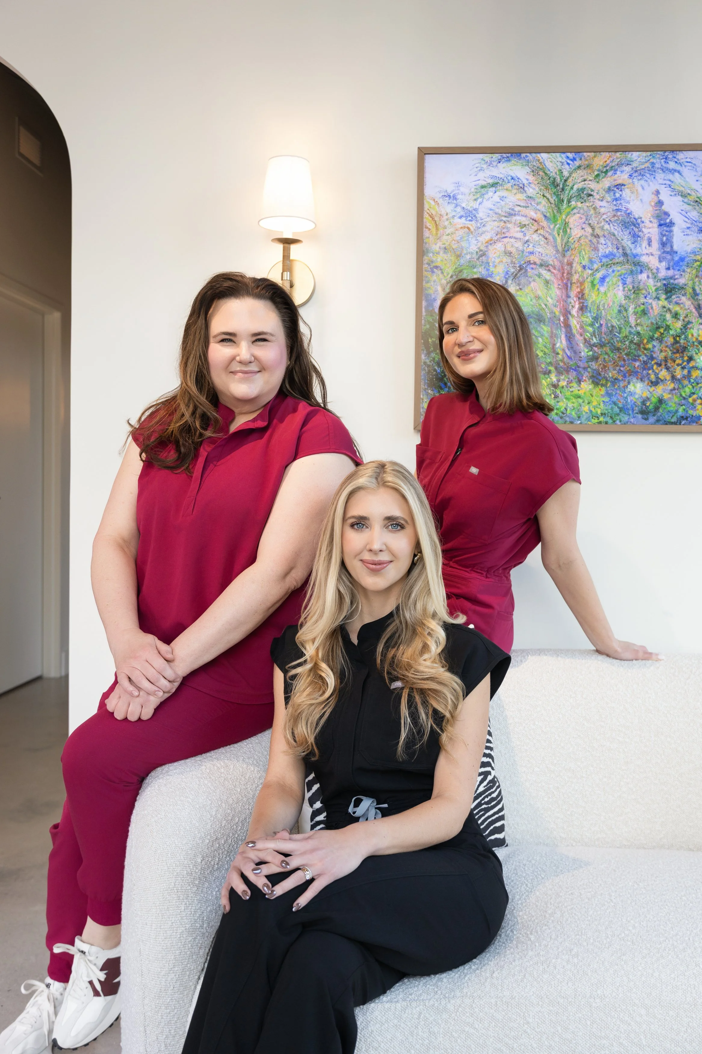 Three women, two standing and one sitting on a white couch, with a colorful abstract painting and a wall sconce in the background.