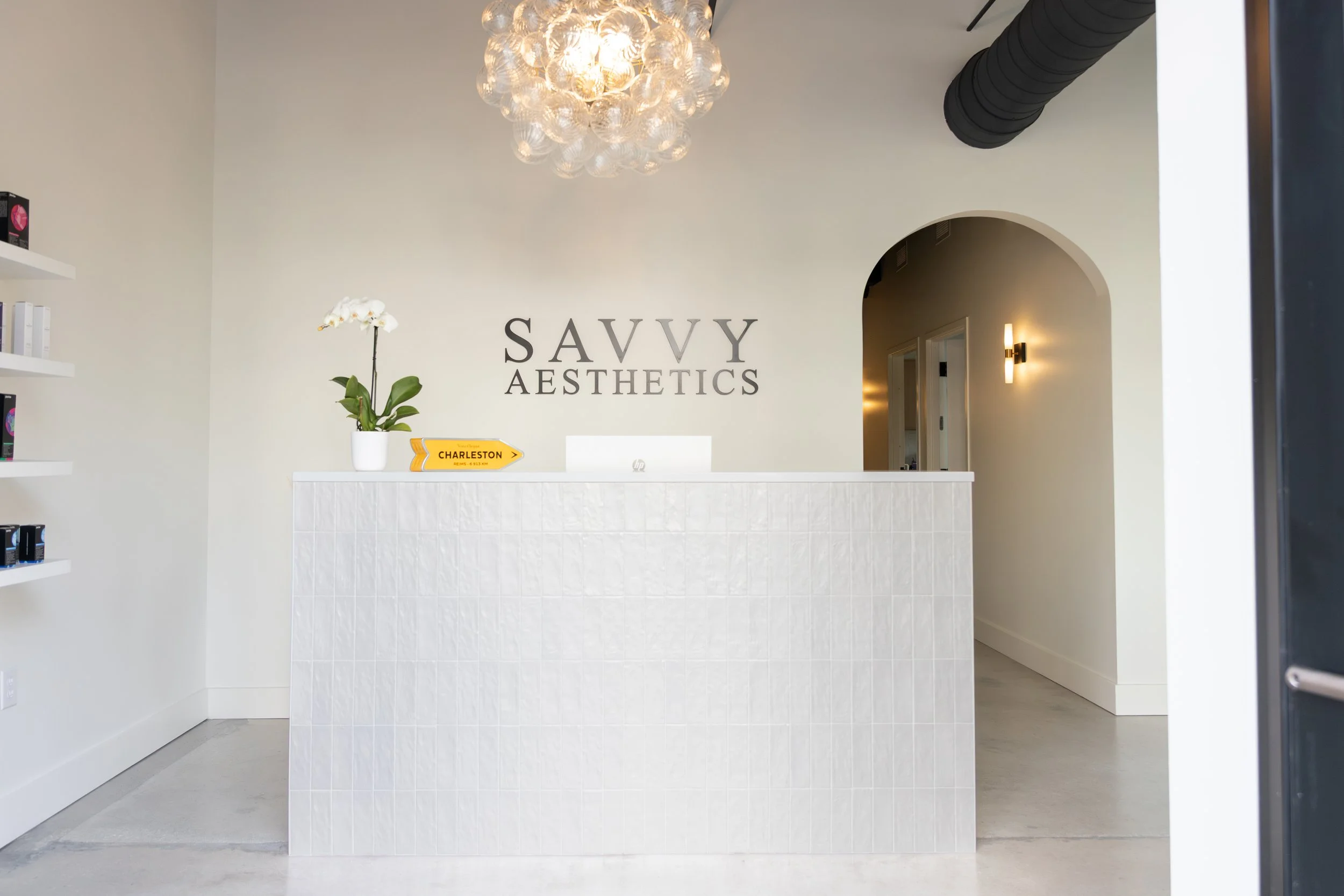 Reception area of Savvy Aesthetics with a white tiled front desk, a potted orchid, and a Charleston sign, with a modern chandelier overhead and a hallway to the right.