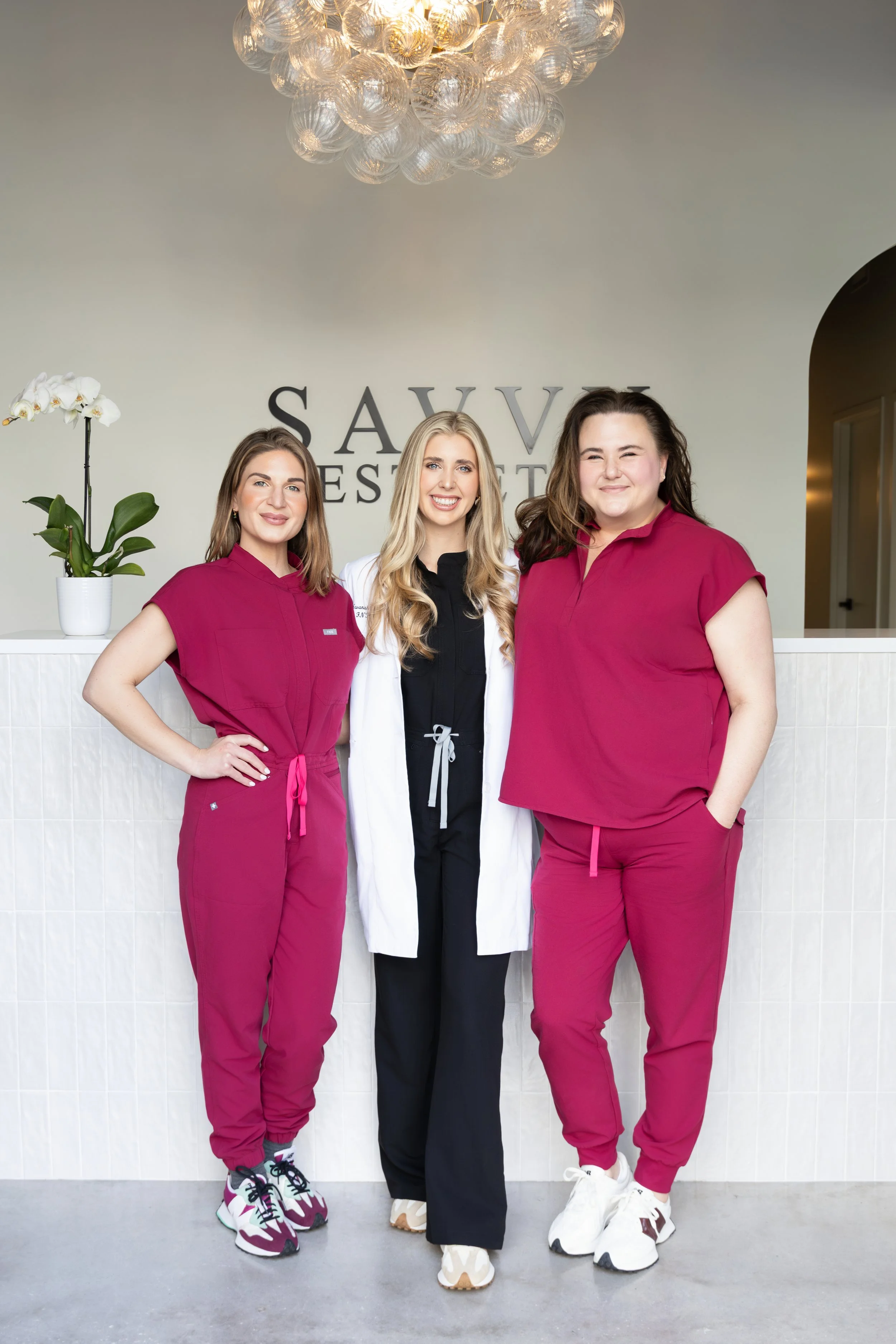 Three women standing in front of a reception desk inside a medical or spa facility, smiling at the camera. The woman on the left is wearing a maroon uniform, the woman in the middle is wearing a white coat, and the woman on the right is wearing a mar