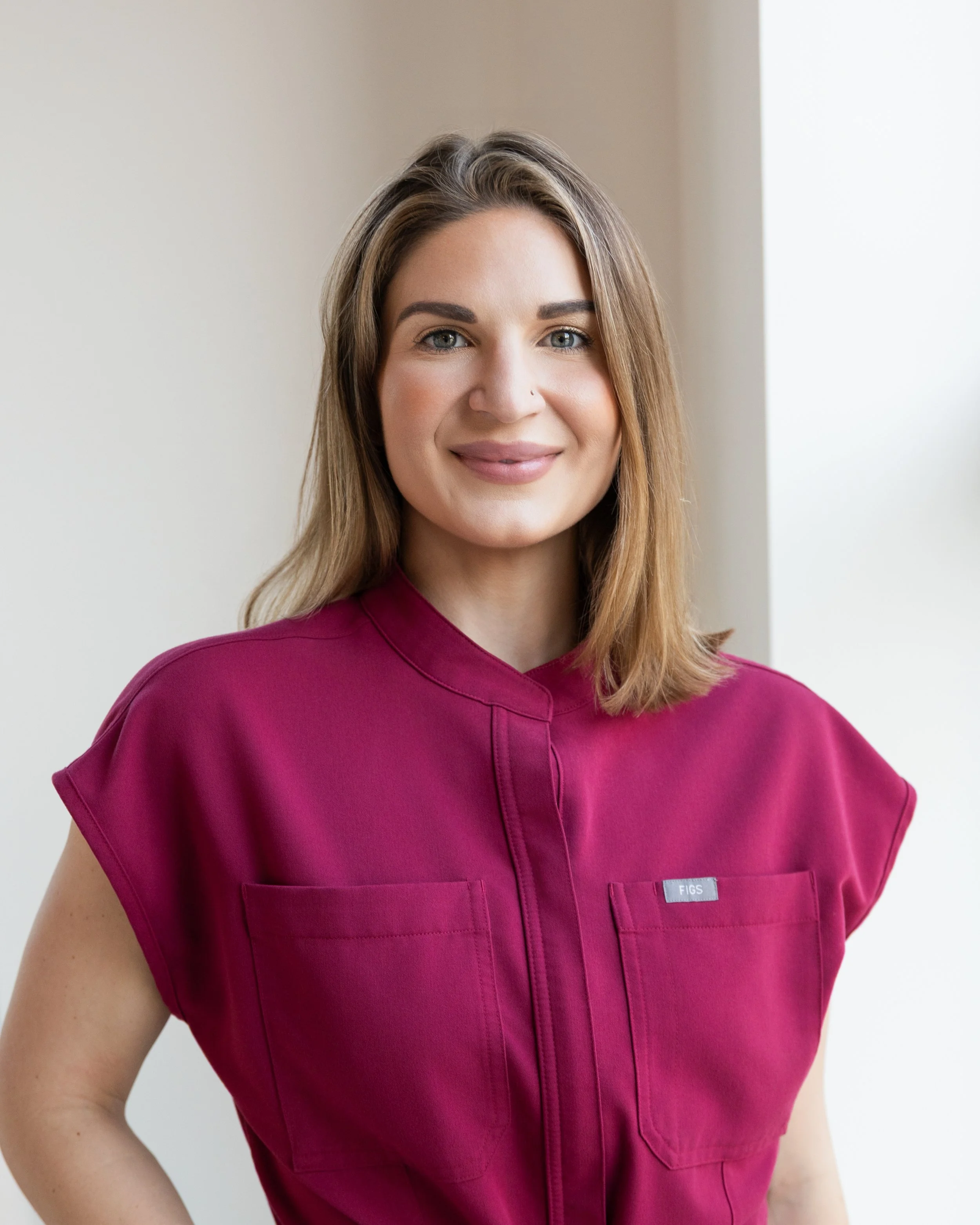 A woman with shoulder-length brown hair, wearing a magenta top, smiling in front of a light-colored wall.