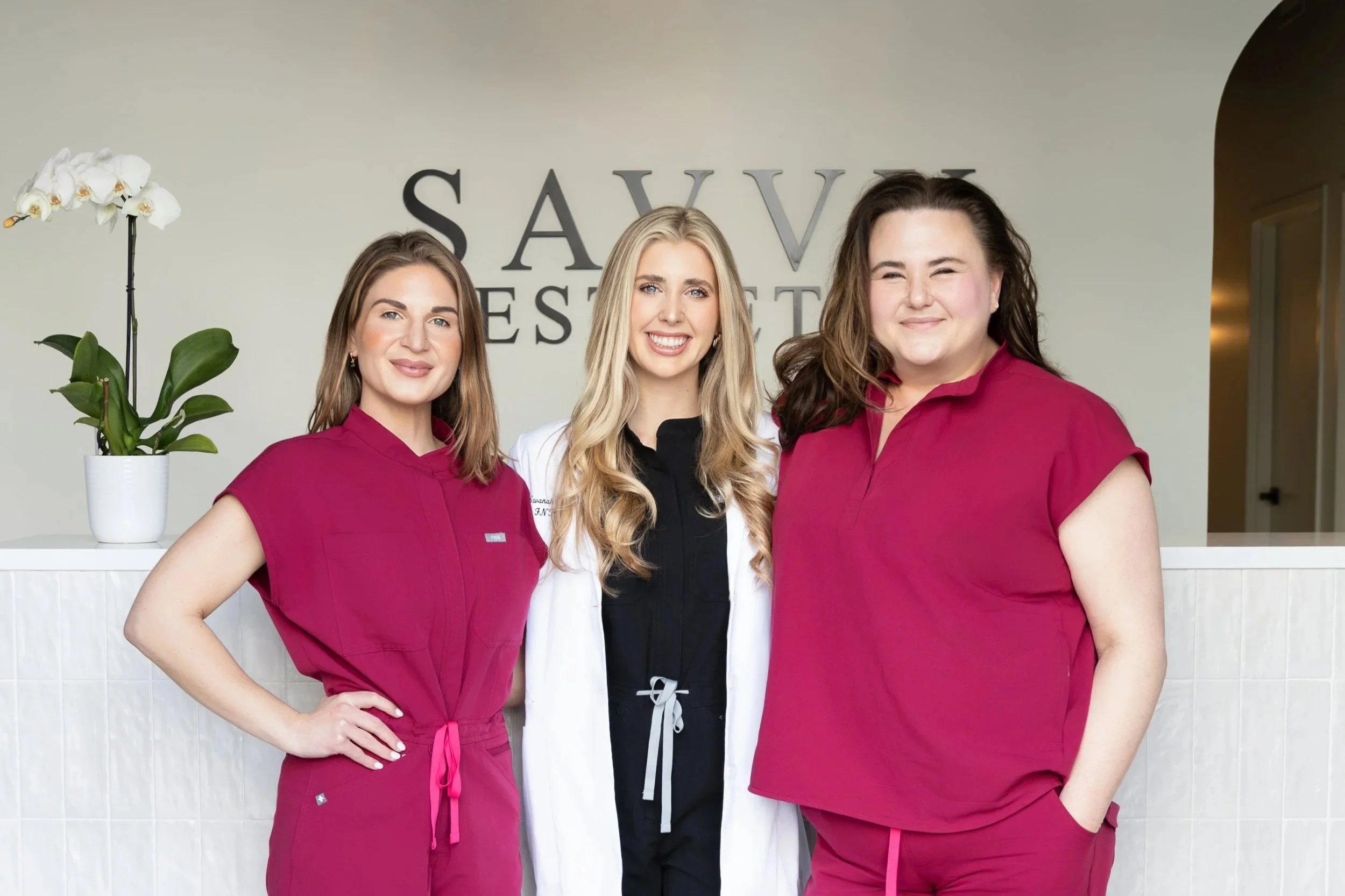 Three women standing together in front of a reception area with a sign that reads 'SAVVY AESTHETICS'