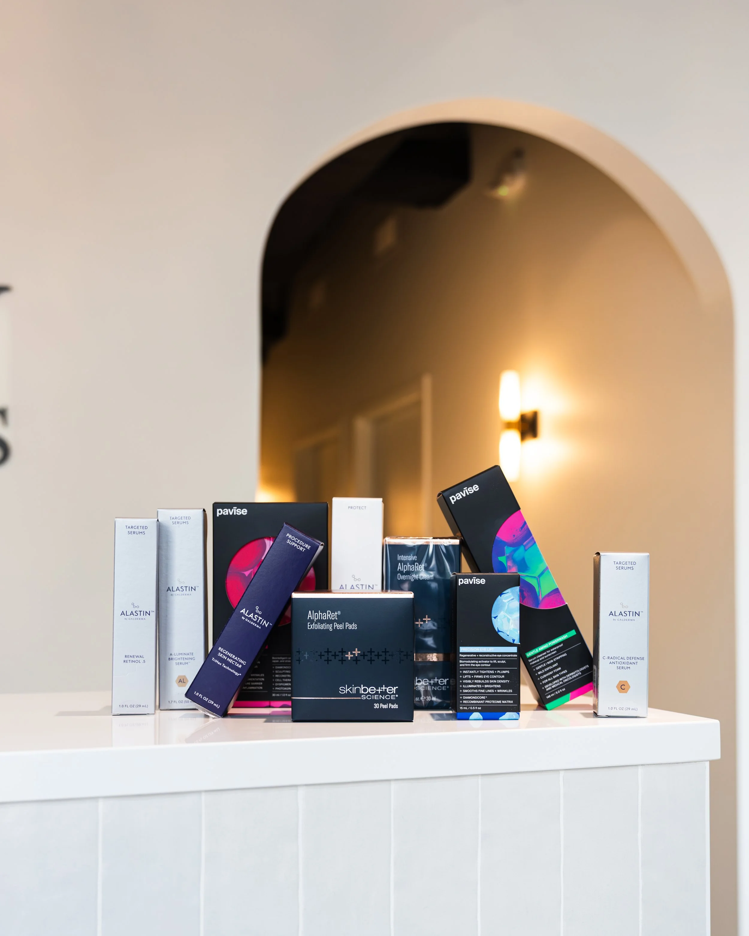 Various skincare and cosmetic products on a white counter in front of a beige wall with arched opening and wall-mounted light fixture.