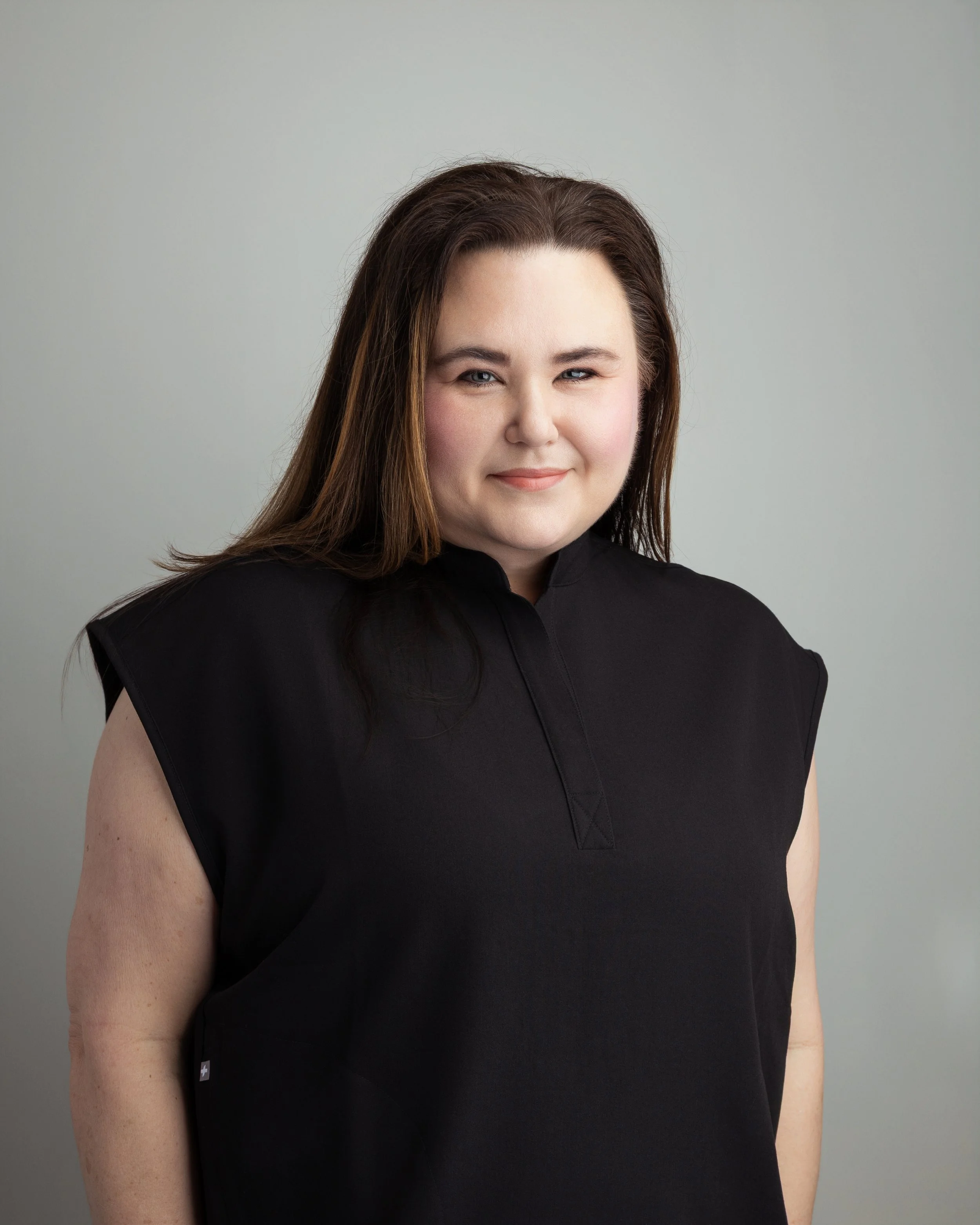 A woman with long brown hair wearing a black sleeveless top, standing against a plain gray background, looking at the camera with a slight smile.