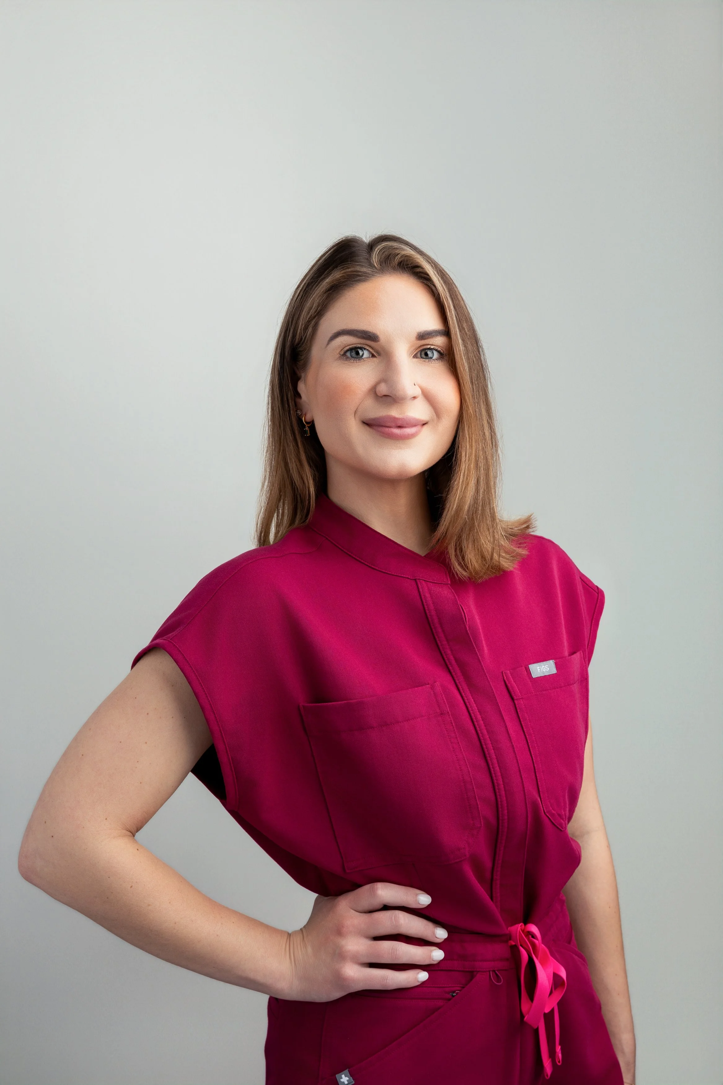 A woman with brown hair and blue eyes wearing a dark pink scrubs top, standing with one hand on her hip and smiling in front of a plain white wall.
