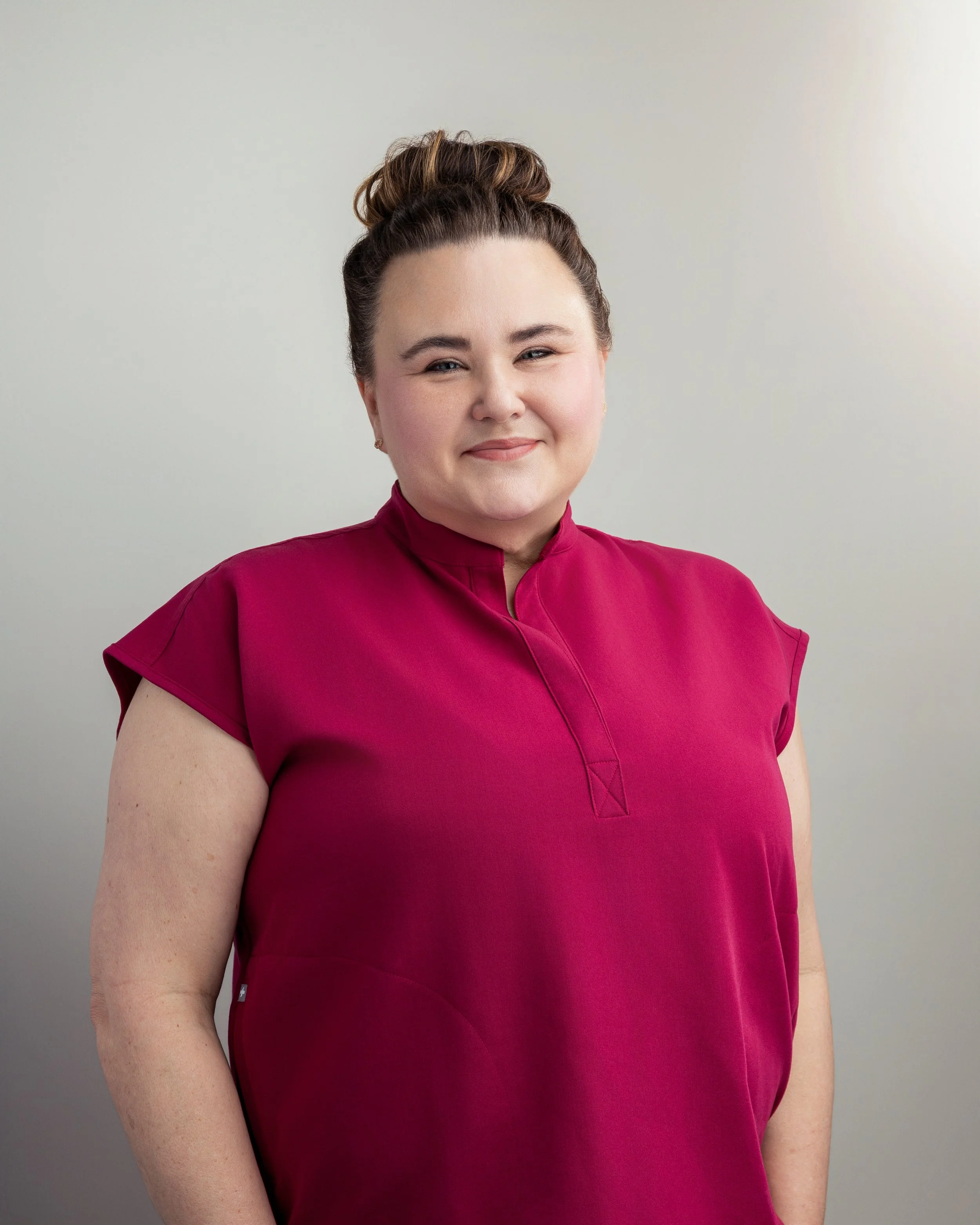 A woman with a bun hairstyle wearing a magenta top, standing against a plain light background, smiling softly.