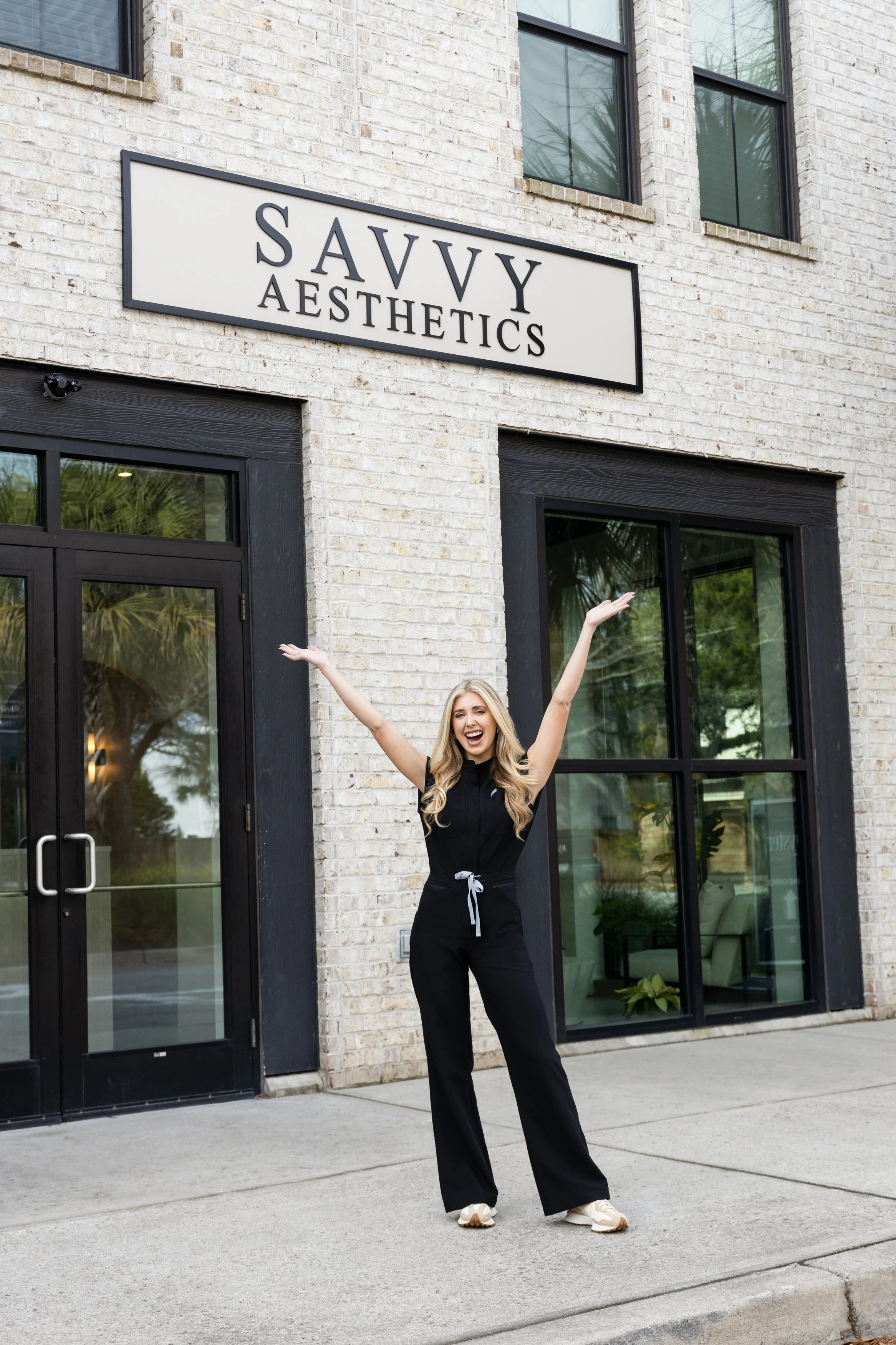 A woman with long blonde hair, wearing a black jumpsuit and white sneakers, standing outside a building called 'Savvy Aesthetics' with her arms raised and smiling.