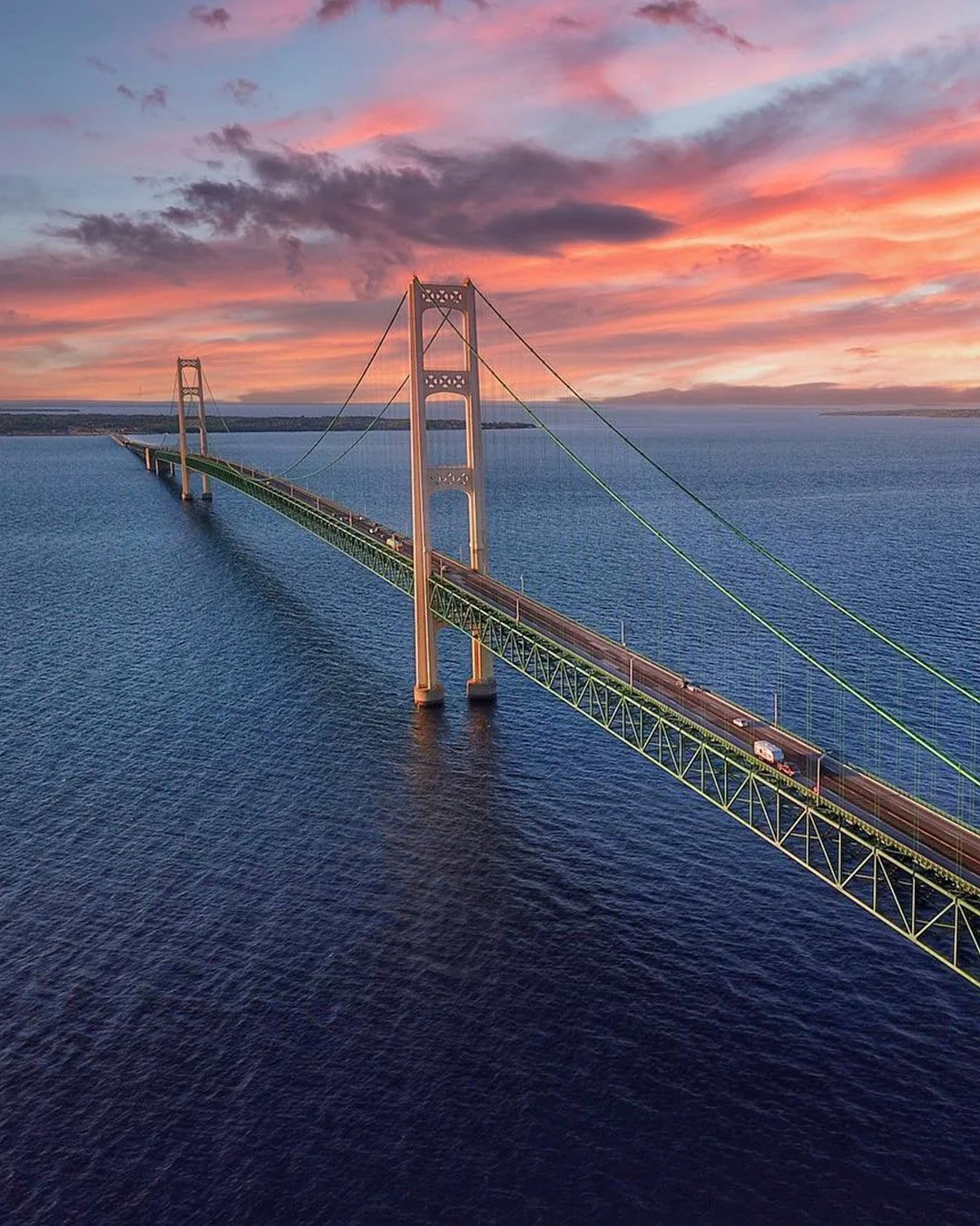 A suspension bridge extending over a body of water at sunset, with pink and purple clouds in the sky.