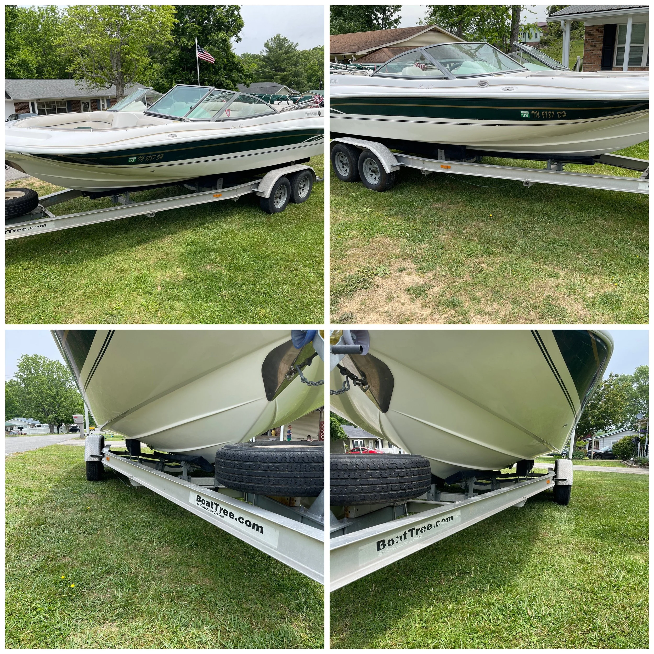 A boat on a trailer parked on a grassy lawn, with a house and trees in the background. The boat is white with green and black accents, and a small American flag is mounted towards the back. The trailer has dual axles and a spare tire mounted on it.