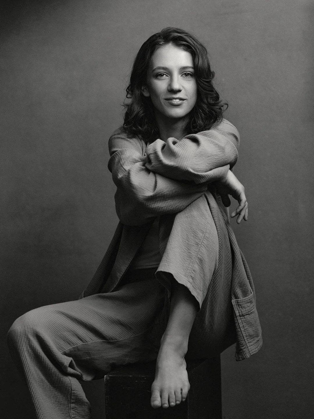A black and white portrait of a woman with shoulder-length wavy hair, sitting on a stool, wearing loose casual clothing, looking at the camera with her arms crossed over her knees, against a plain background.