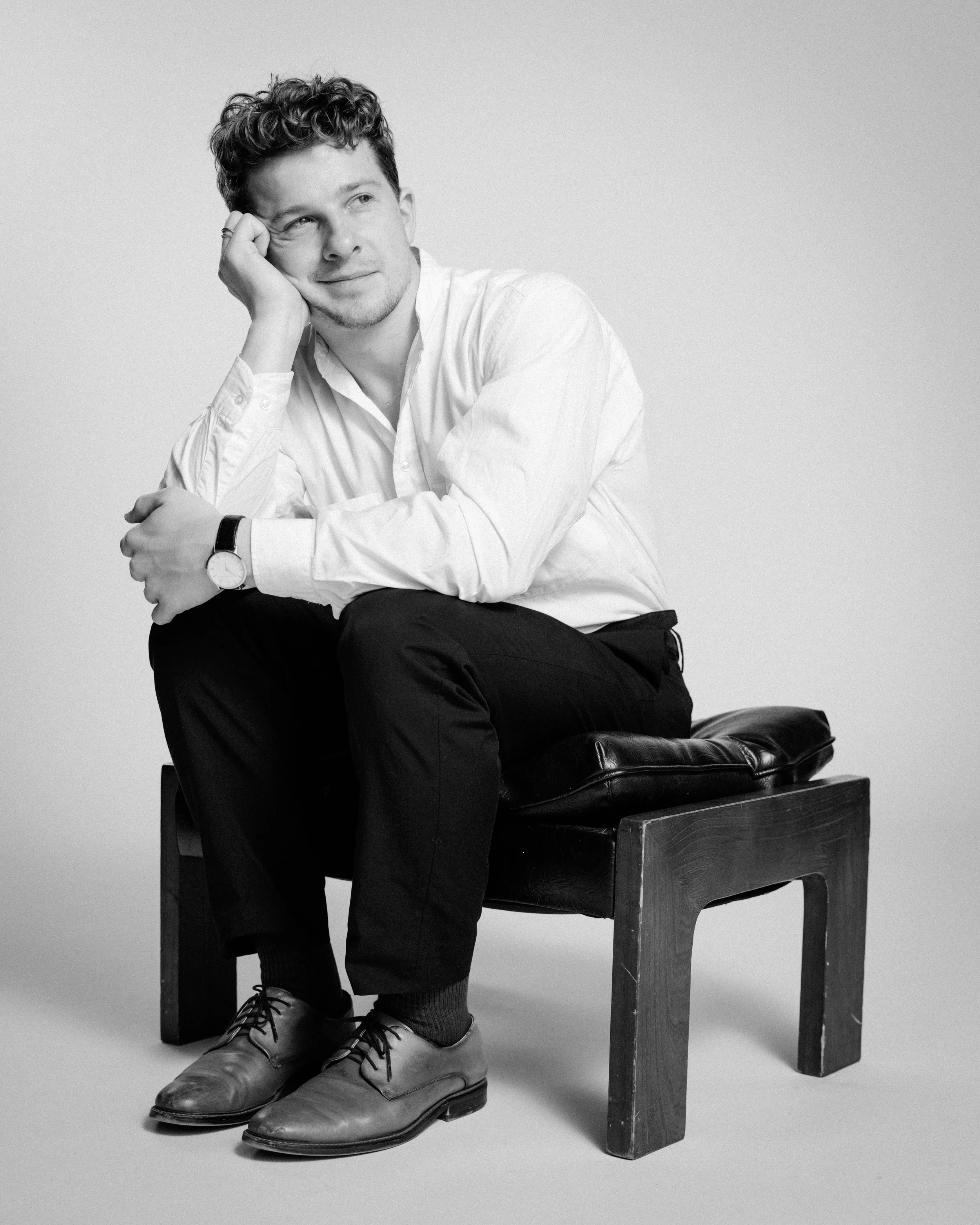 Black and white portrait of a young man sitting on a wooden stool with a cushioned top. He is wearing a white shirt, dark pants, and dress shoes. He has curly hair and is resting his head on his left hand, with his right arm resting on his knee. He is looking thoughtfully to the side.