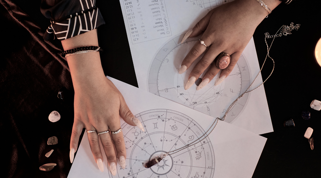 Hands of a woman with rings and nails, pointing at astrology charts on a black surface, surrounded by gemstones and jewelry.