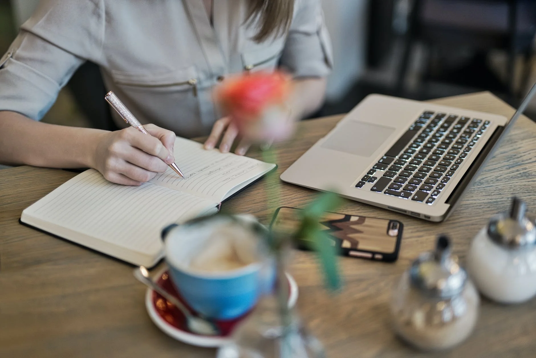 A woman takes notes in a notebook at a wooden table with a laptop, a phone, and cups of coffee, with decorative sugar containers nearby.