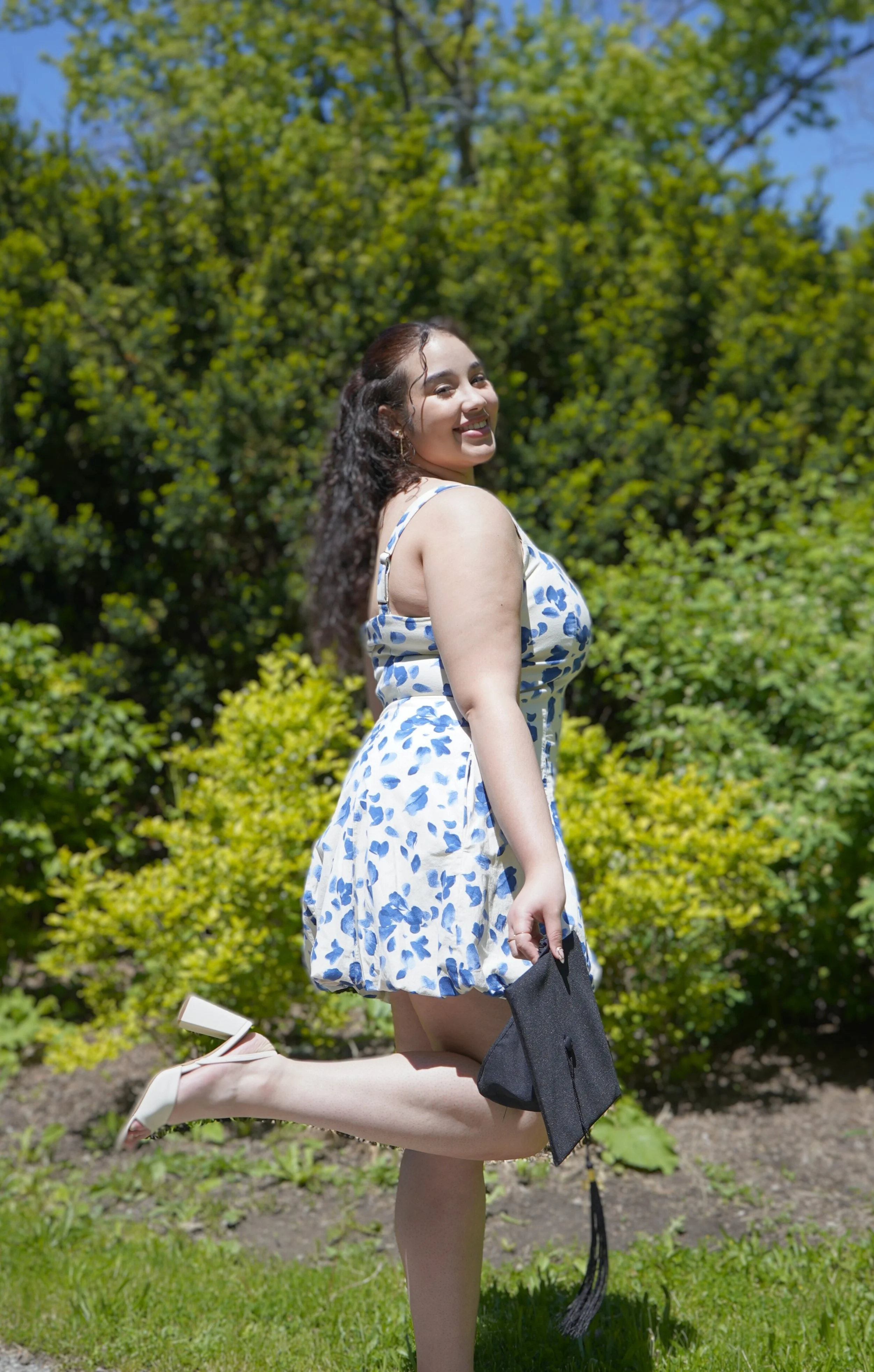 A young woman in a floral dress holding a graduation cap, smiling outdoors on a sunny day with green bushes and trees in the background.