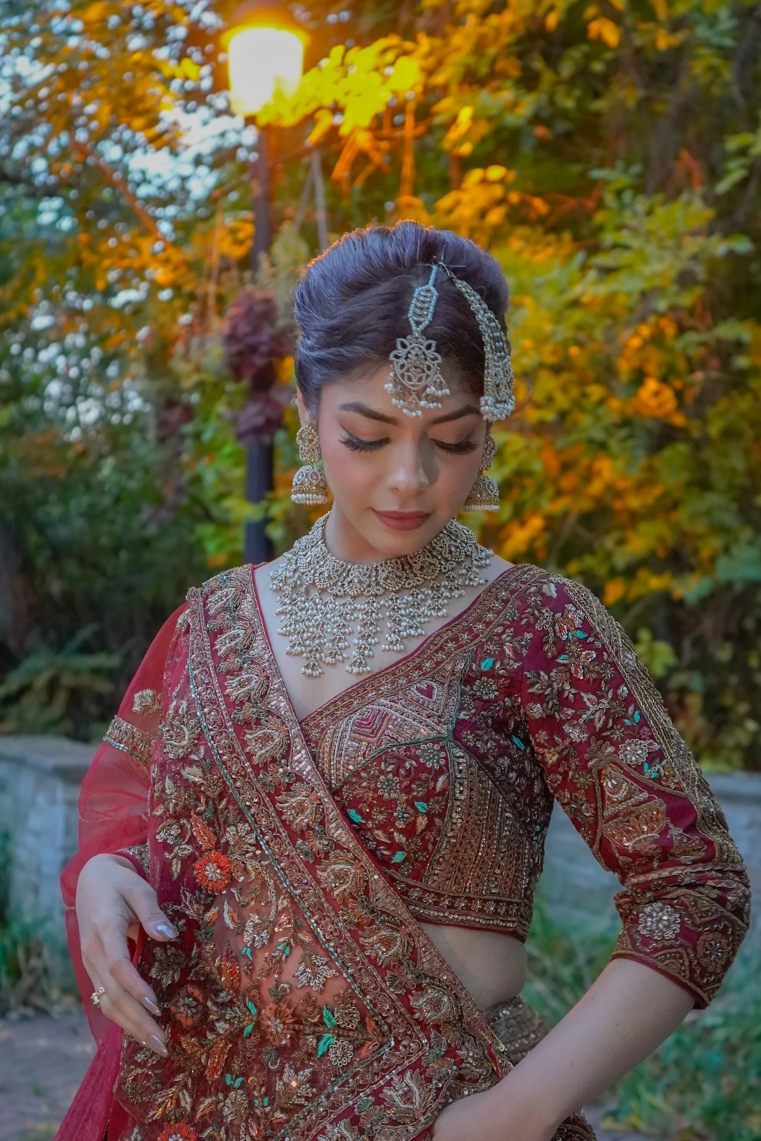 A woman dressed in traditional Indian bridal attire, wearing intricate jewelry, a richly embroidered red saree, and standing outdoors with autumn-colored foliage in the background.