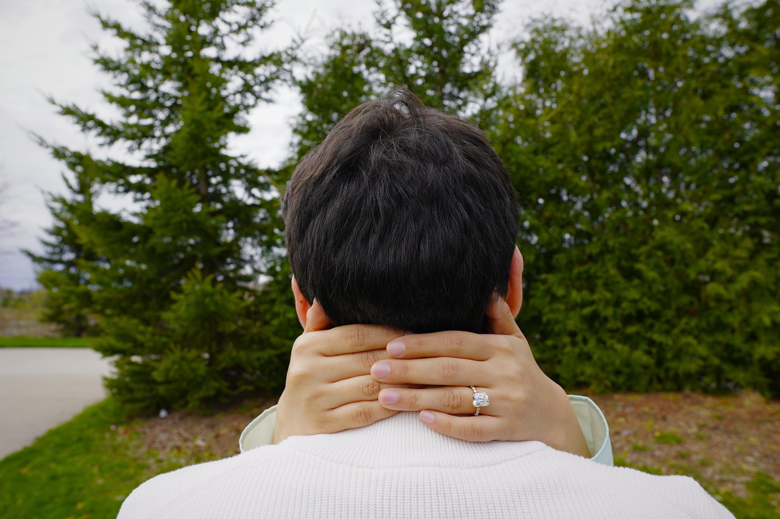 A woman with a engagement ring on her finger holds her hands around a man’s neck from behind outdoors, with green trees in the background.