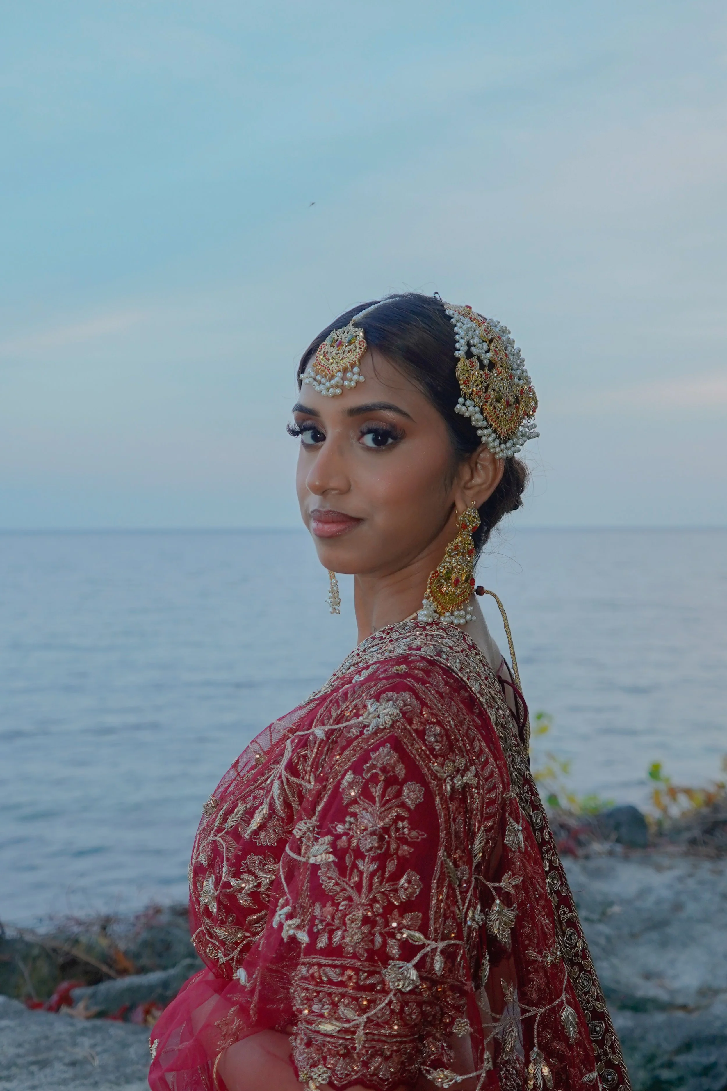 A woman dressed in traditional Indian attire and jewelry standing outdoors by the water during evening.