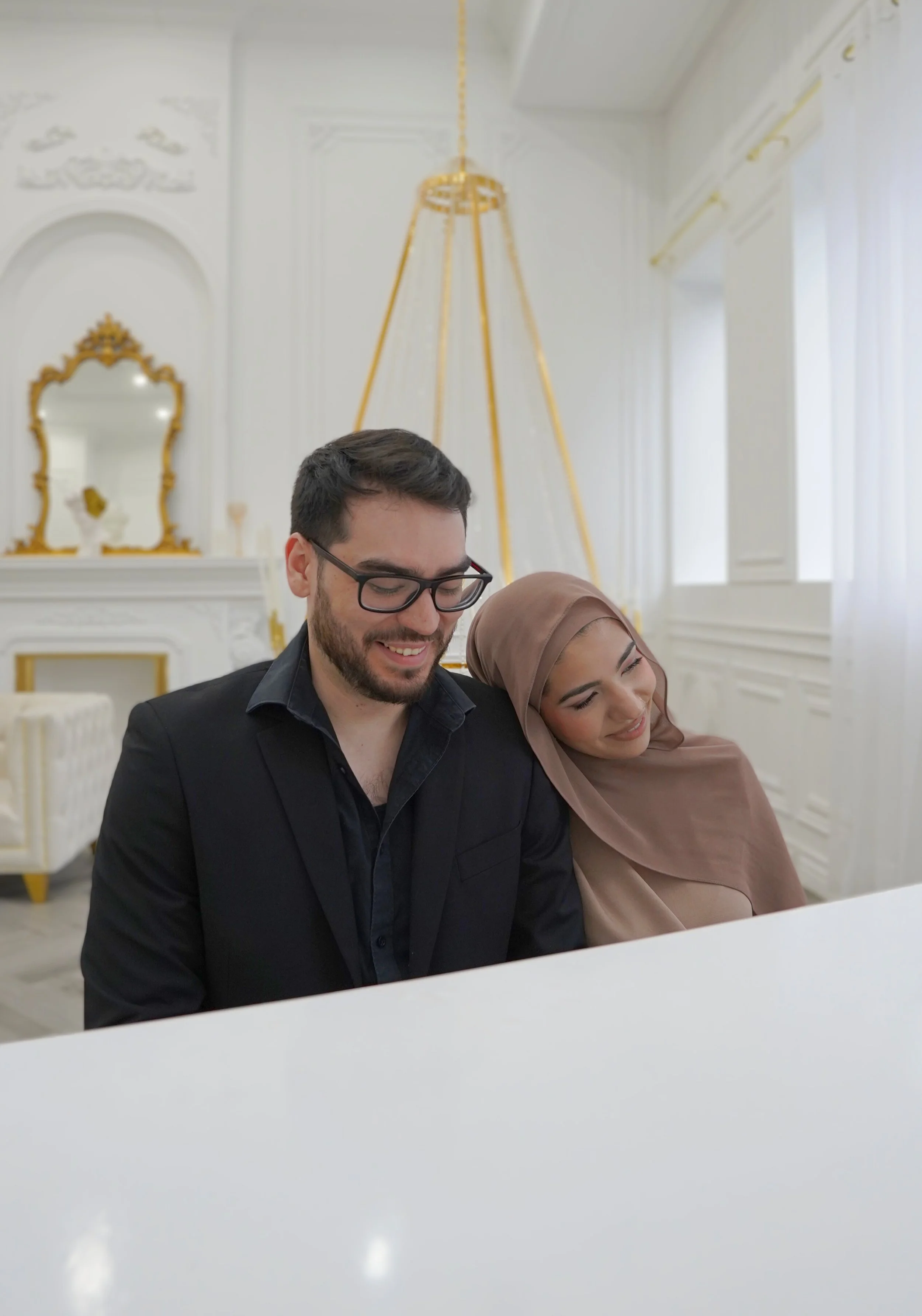 A man and woman sitting together at a white piano, smiling and leaning towards each other in a bright, elegant room with white walls and gold decor.