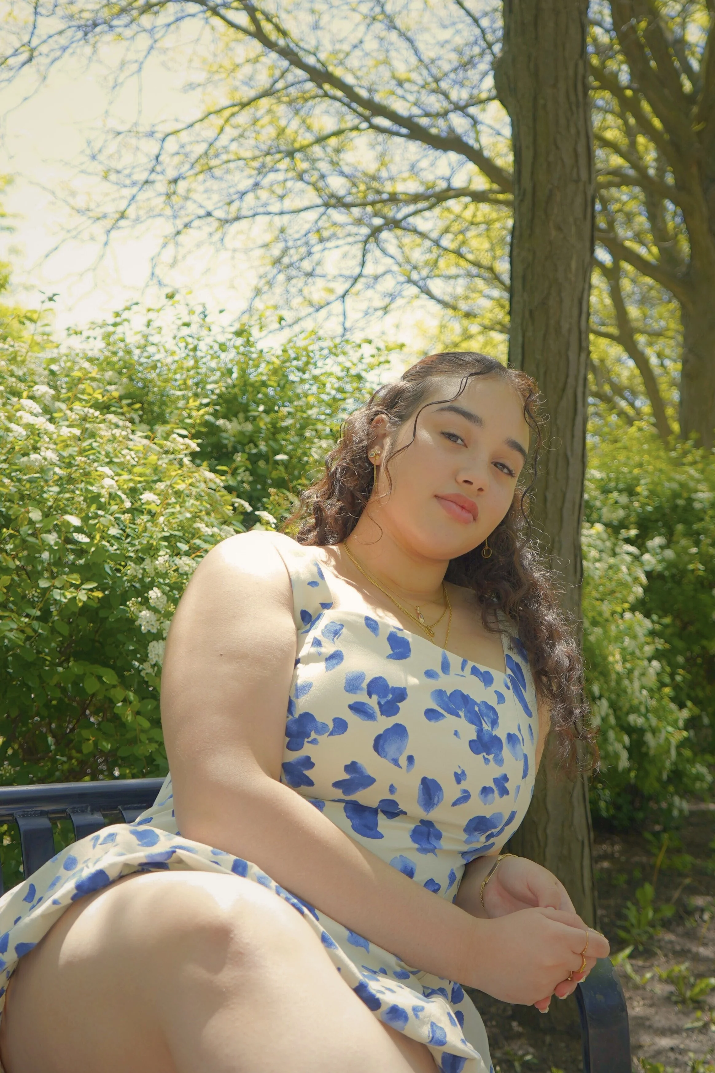 Young woman sitting on a black outdoor bench, wearing a white dress with blue floral pattern, surrounded by green bushes and tall trees on a sunny day.