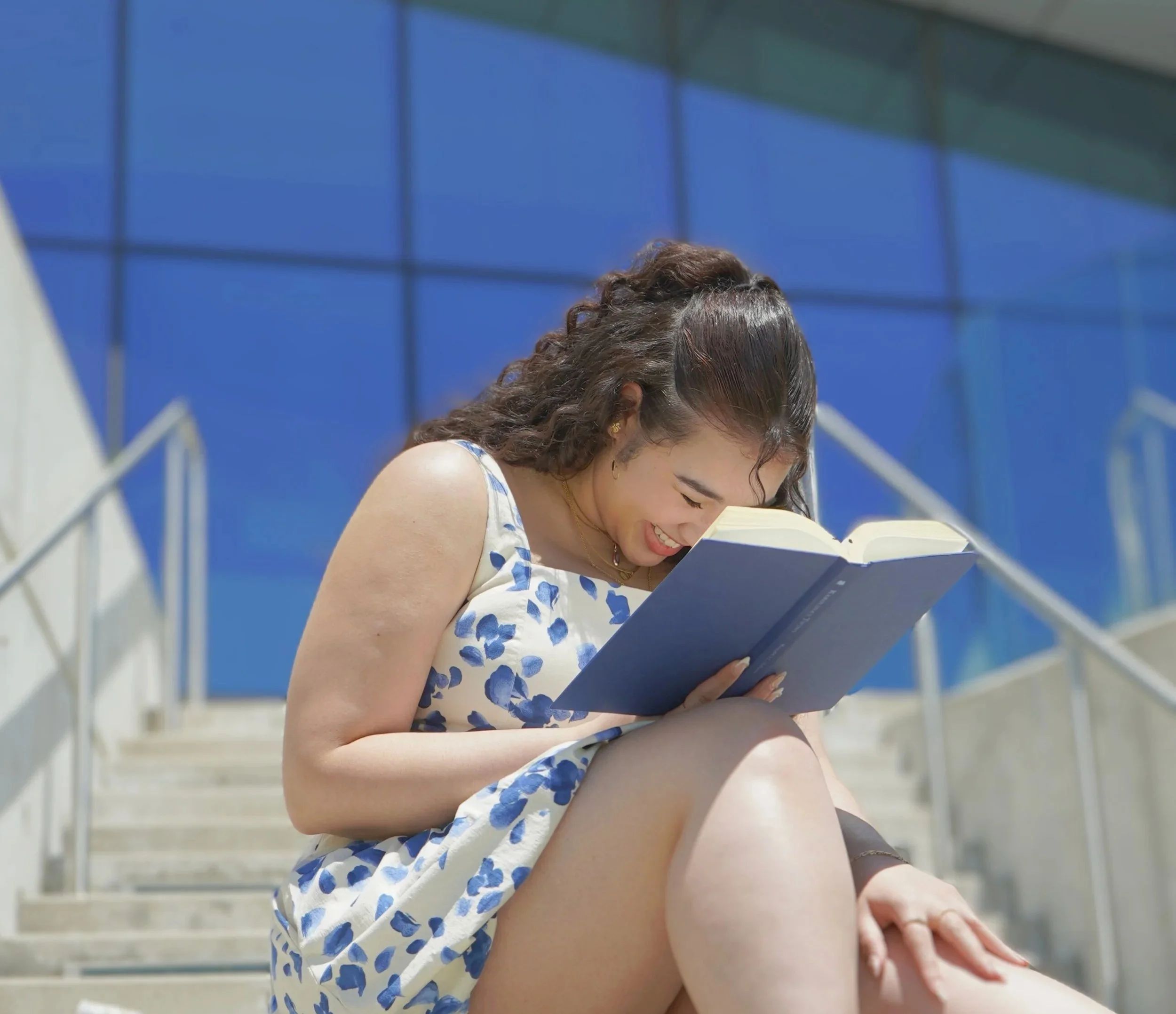 A woman with curly hair sitting on outdoor stairs, smiling and reading a book, against a backdrop of a modern glass building.