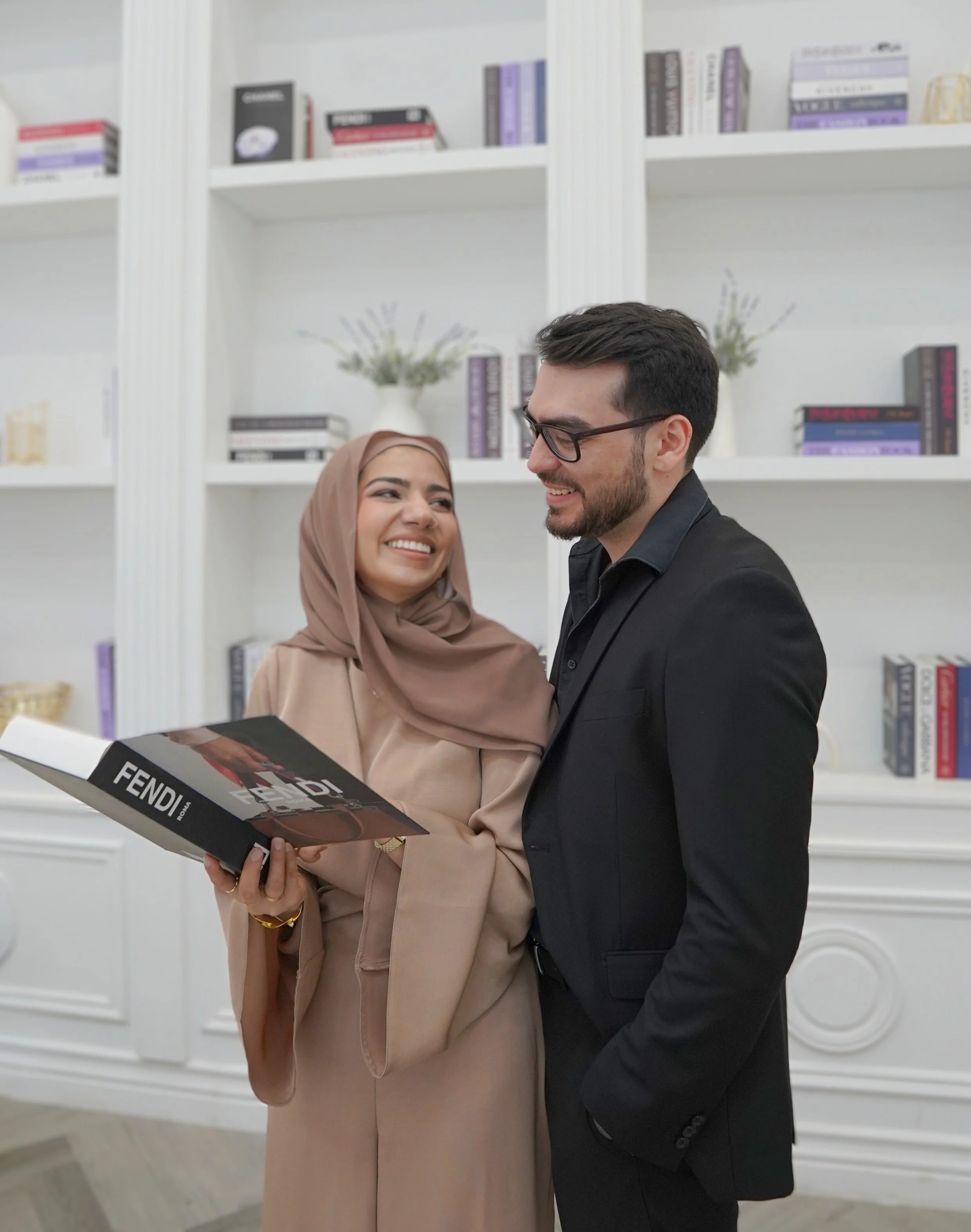 A woman in a beige dress and hijab smiling while holding a Fendi magazine, talking to a man in a black suit and glasses, standing in front of white bookshelves with books and decorations.