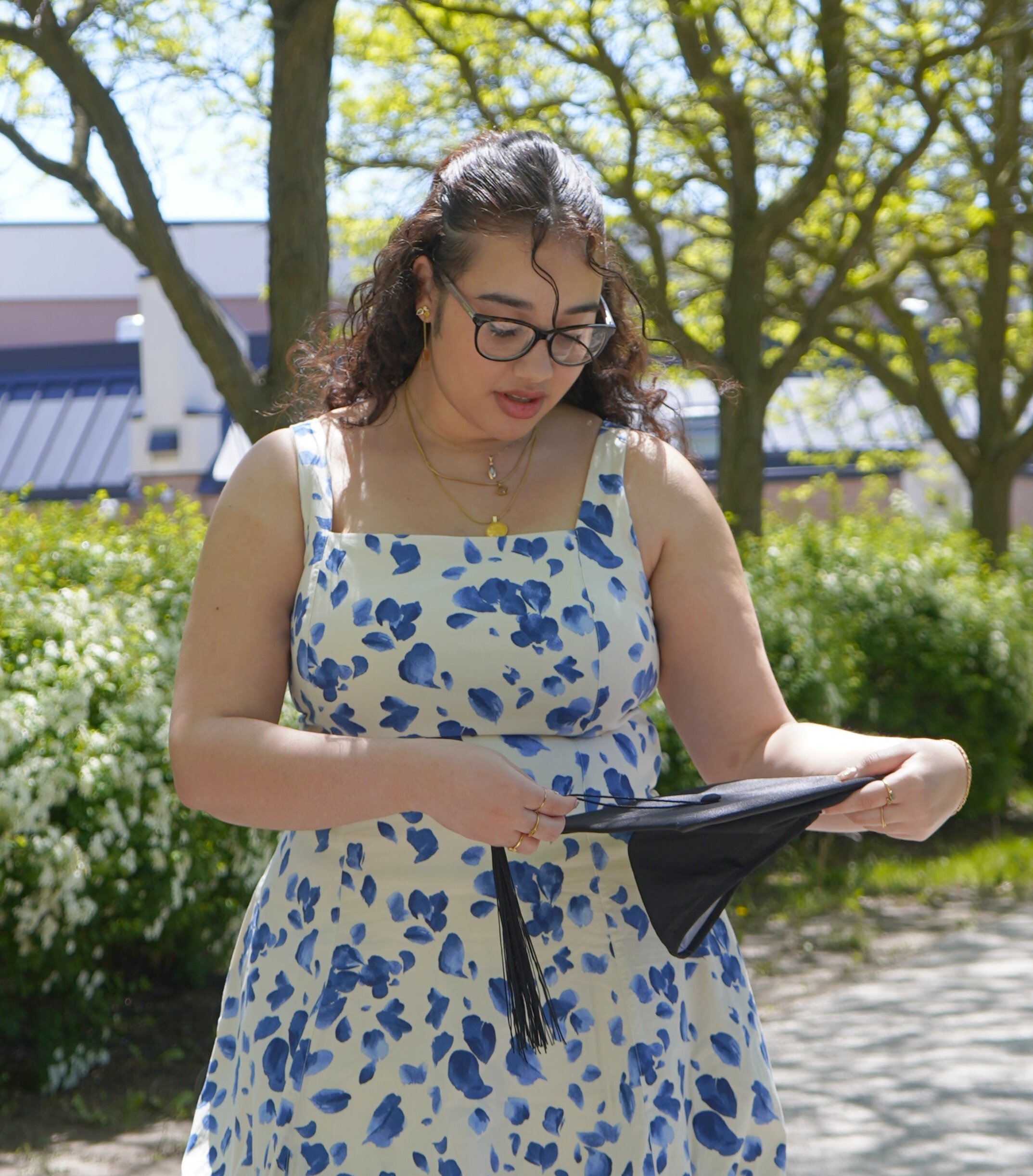 Young woman outdoors wearing glasses and a floral dress holding a diploma or certificate, celebrating a graduation.