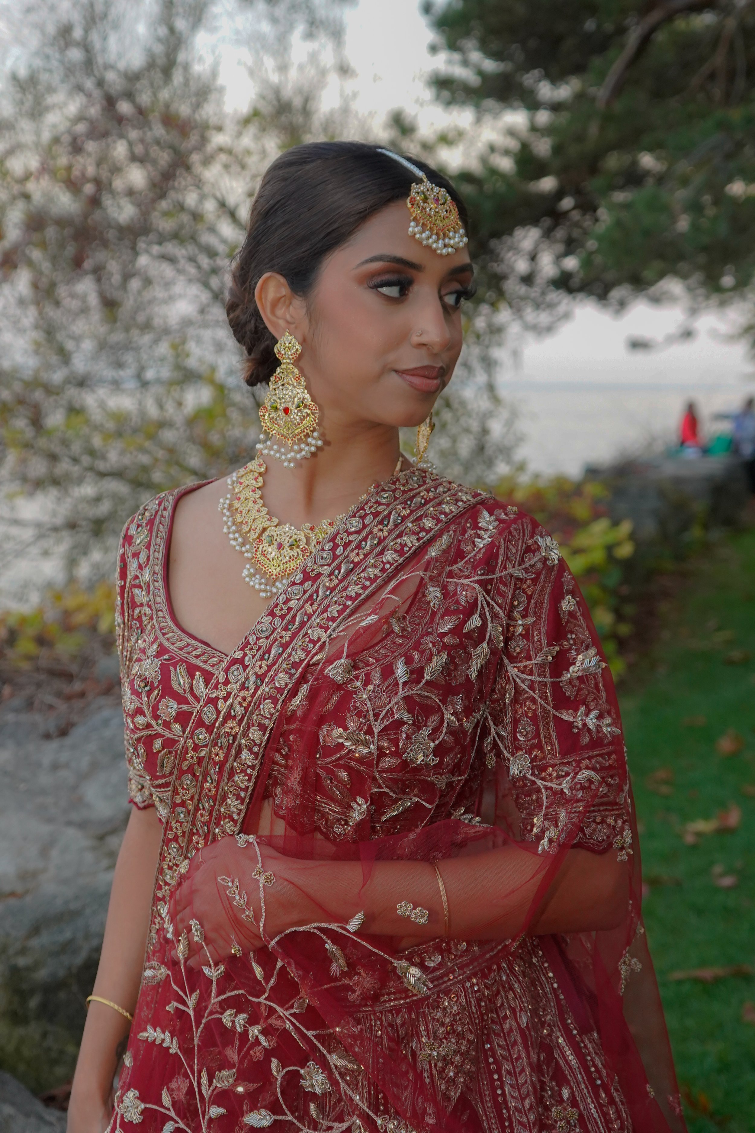 A woman dressed in traditional Indian attire, wearing a red embroidered saree with gold and silver details, and ornate gold jewelry, standing outdoors near a lake or river with trees in the background.