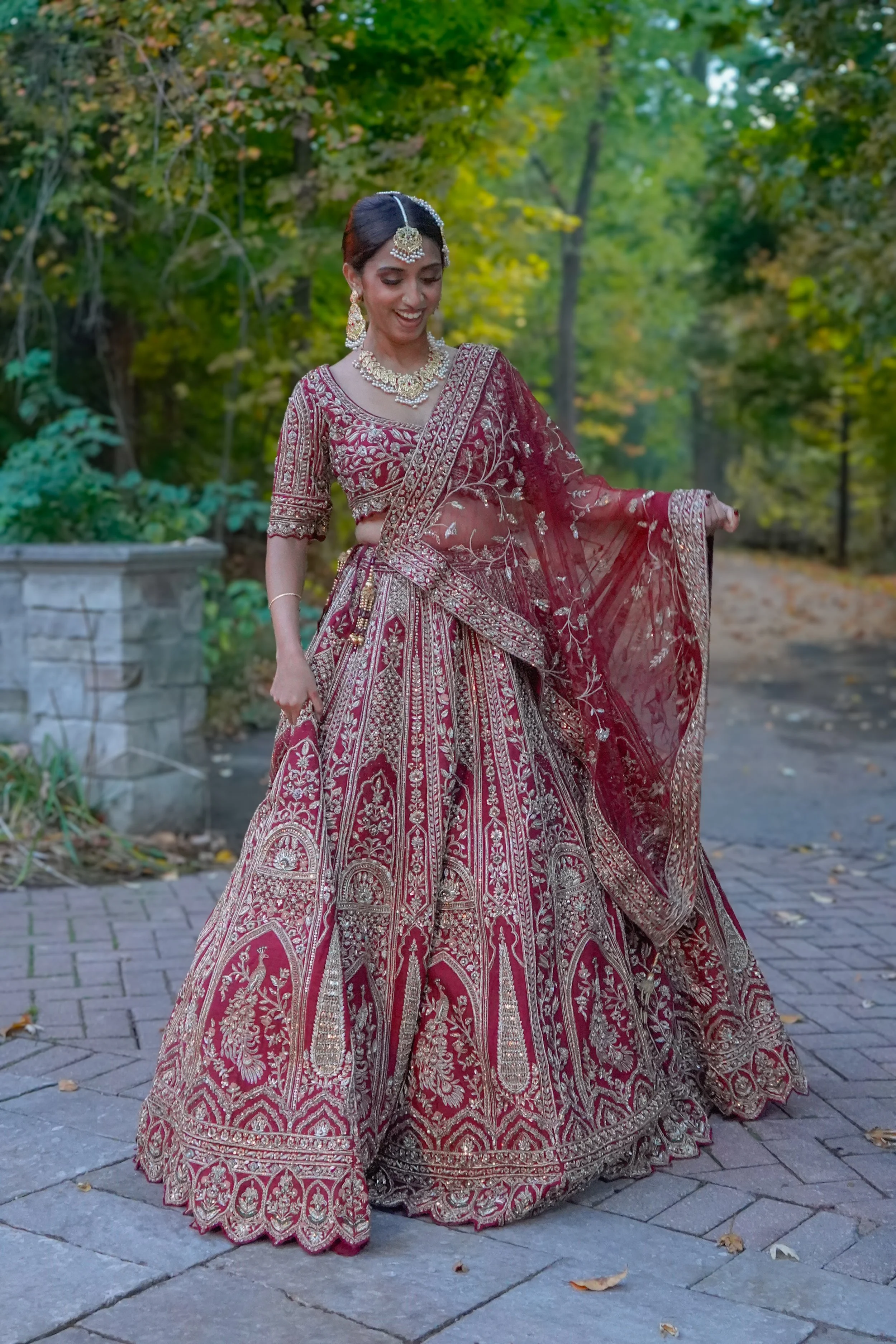 A woman wearing a traditional Indian bridal outfit in maroon and gold, standing on a brick pathway in a wooded area.