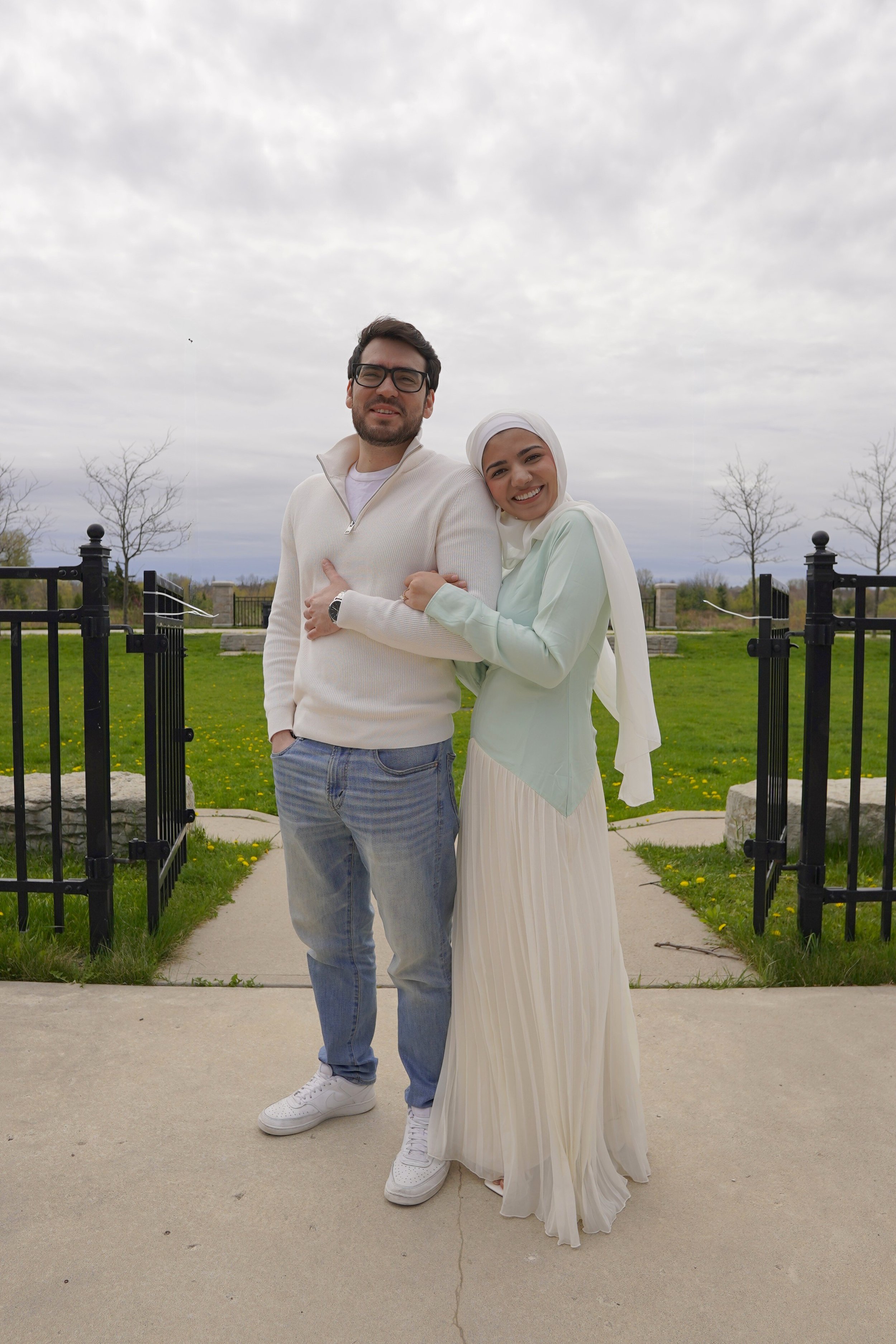 A smiling couple stands together outdoors on a cloudy day, with a woman wearing a white headscarf and a long white and mint green dress, and a man wearing glasses, a white sweater, and jeans, holding hands in front of a park gate and green lawn.