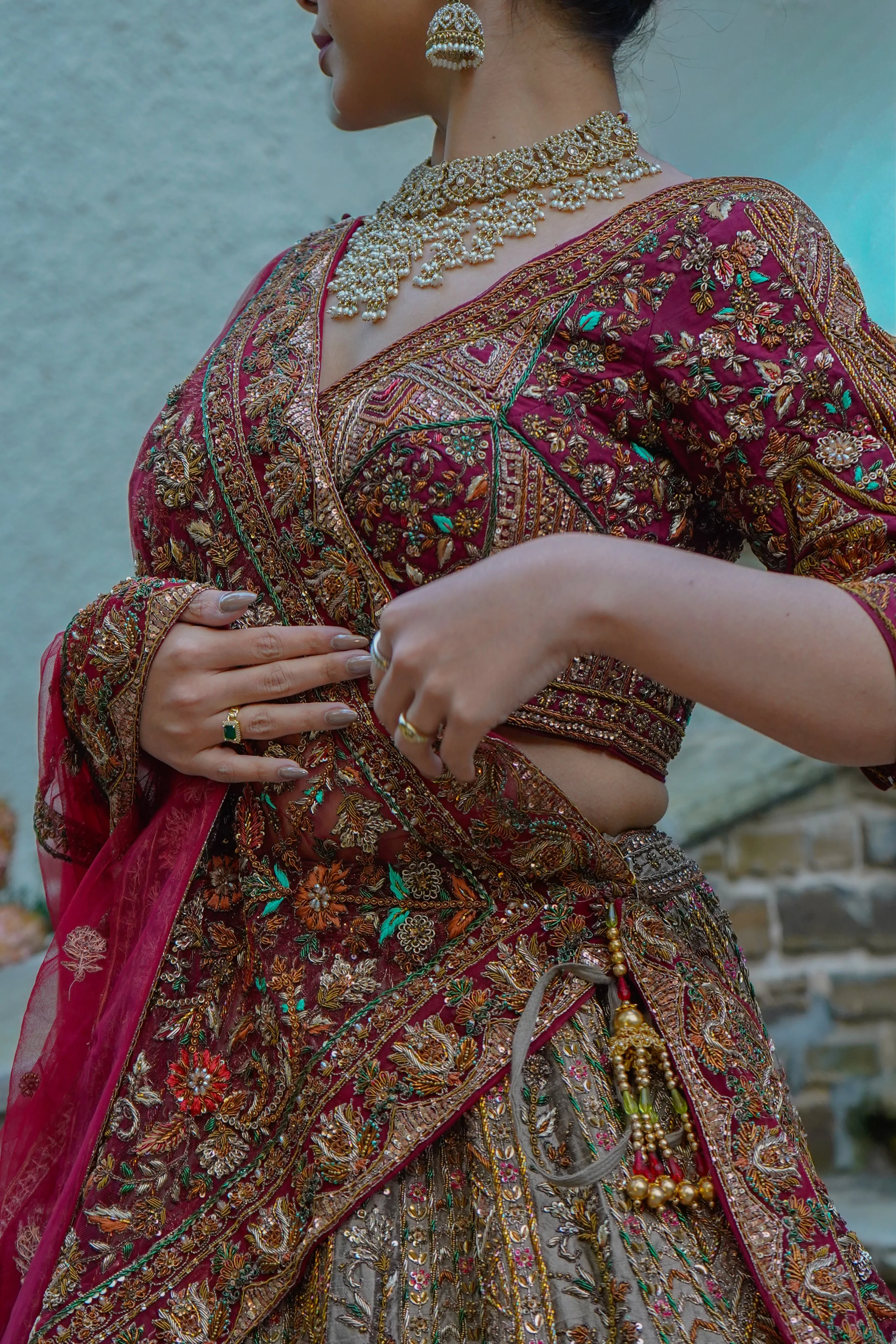 Close-up of a woman wearing traditional, richly embroidered maroon and gold Indian saree with jewelry, including a pearl necklace, earrings, and rings, holding the saree pleats.