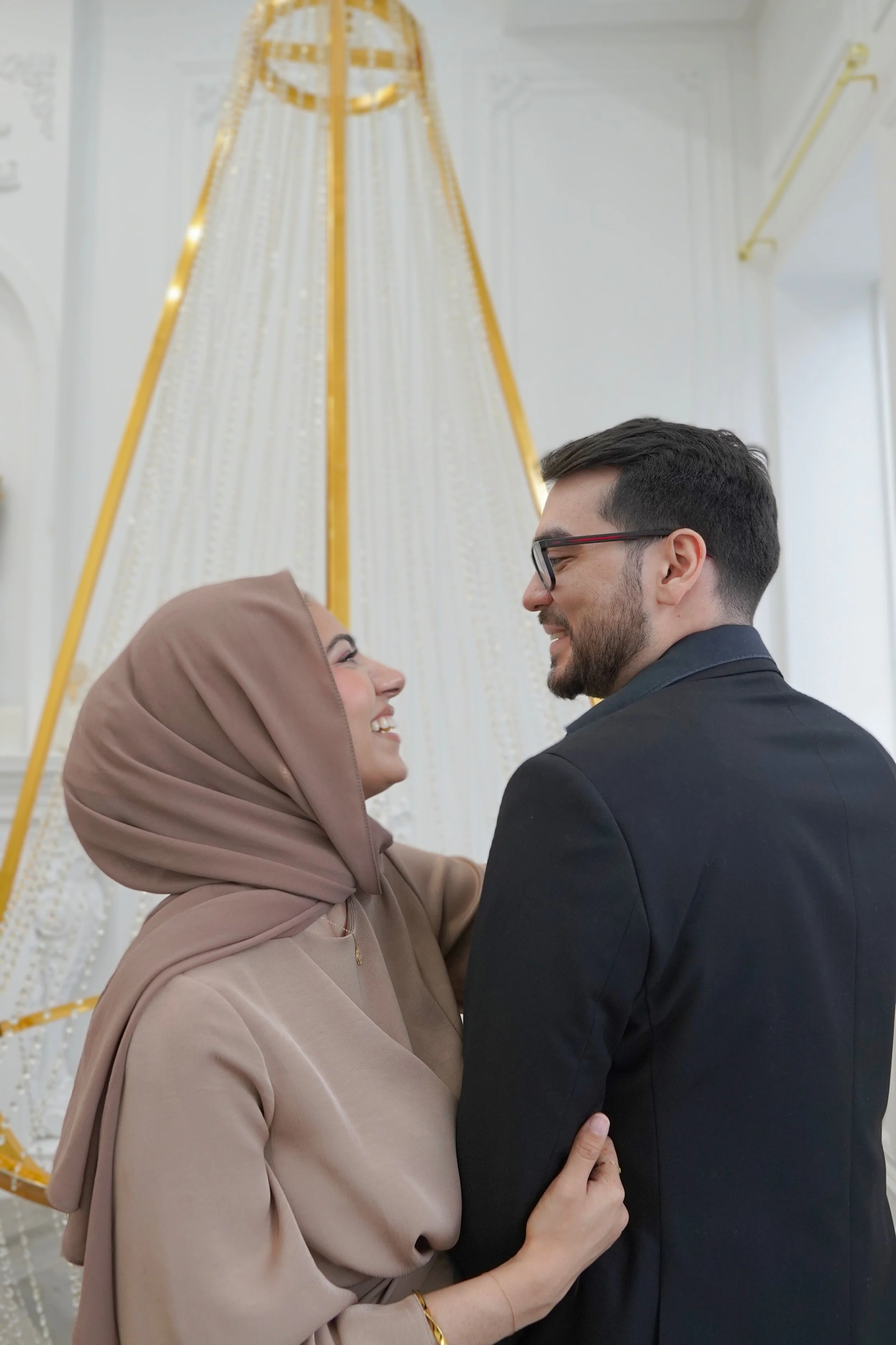 A happy woman in a beige hijab and dress smiling at a man in a black suit with glasses, both indoors with a large ornate chandelier in the background.