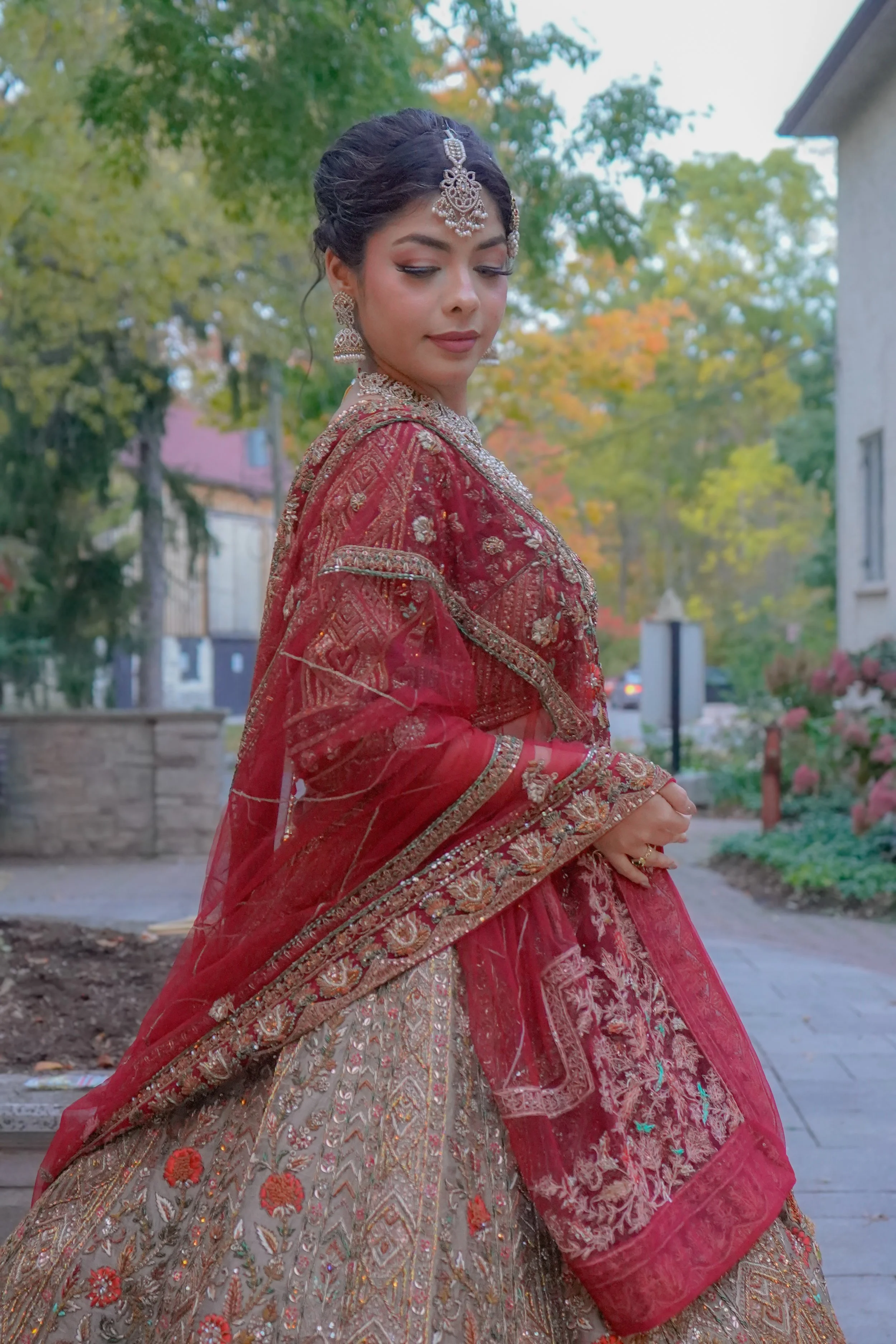 A woman dressed in traditional South Asian bridal attire, wearing a red embroidered lehenga with gold and silver embellishments, along with matching jewelry and a jeweled headpiece, standing outdoors with trees and buildings in the background.