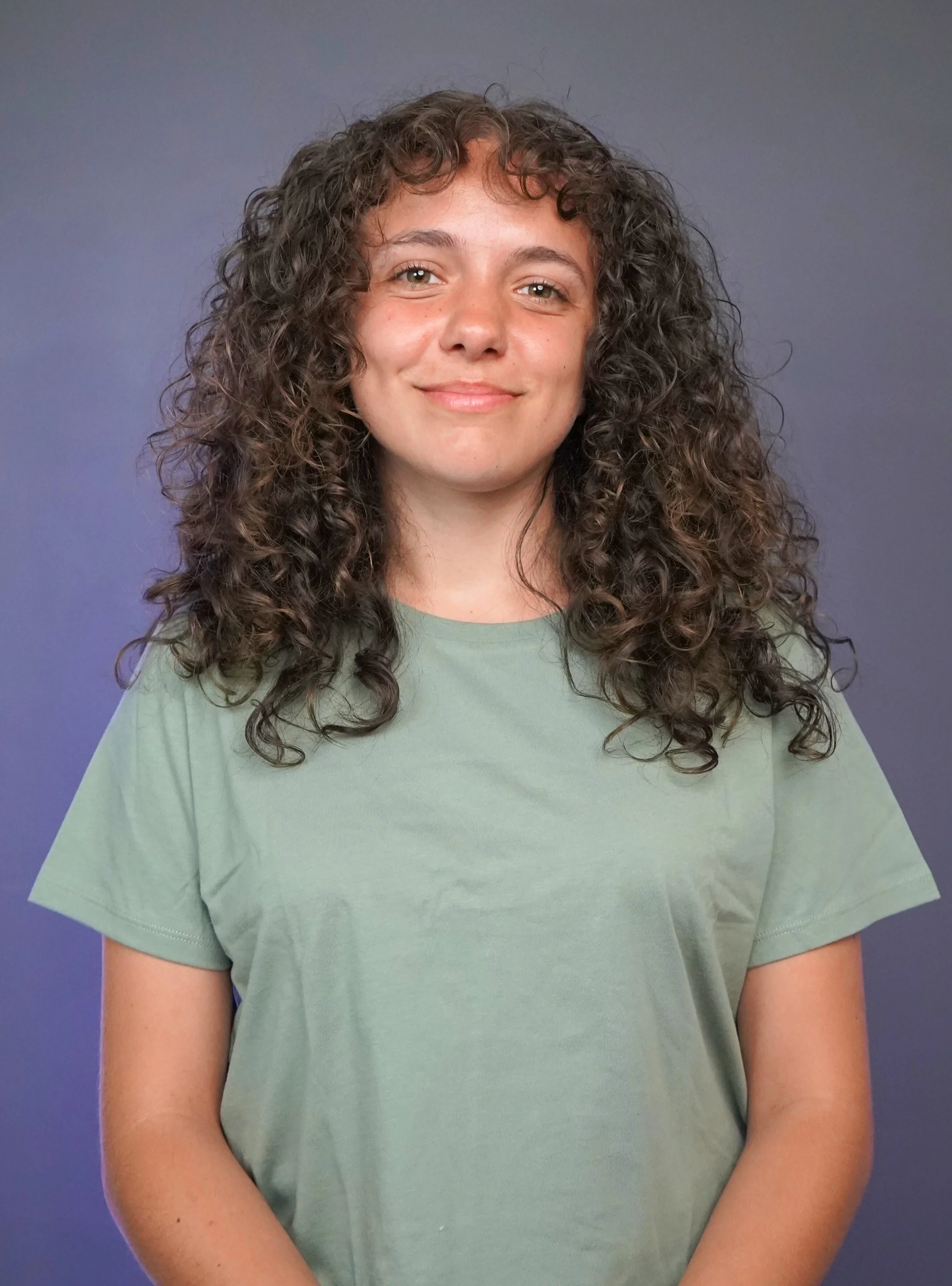 A woman with curly brown hair wearing a light green t-shirt, smiling at the camera against a purple background.