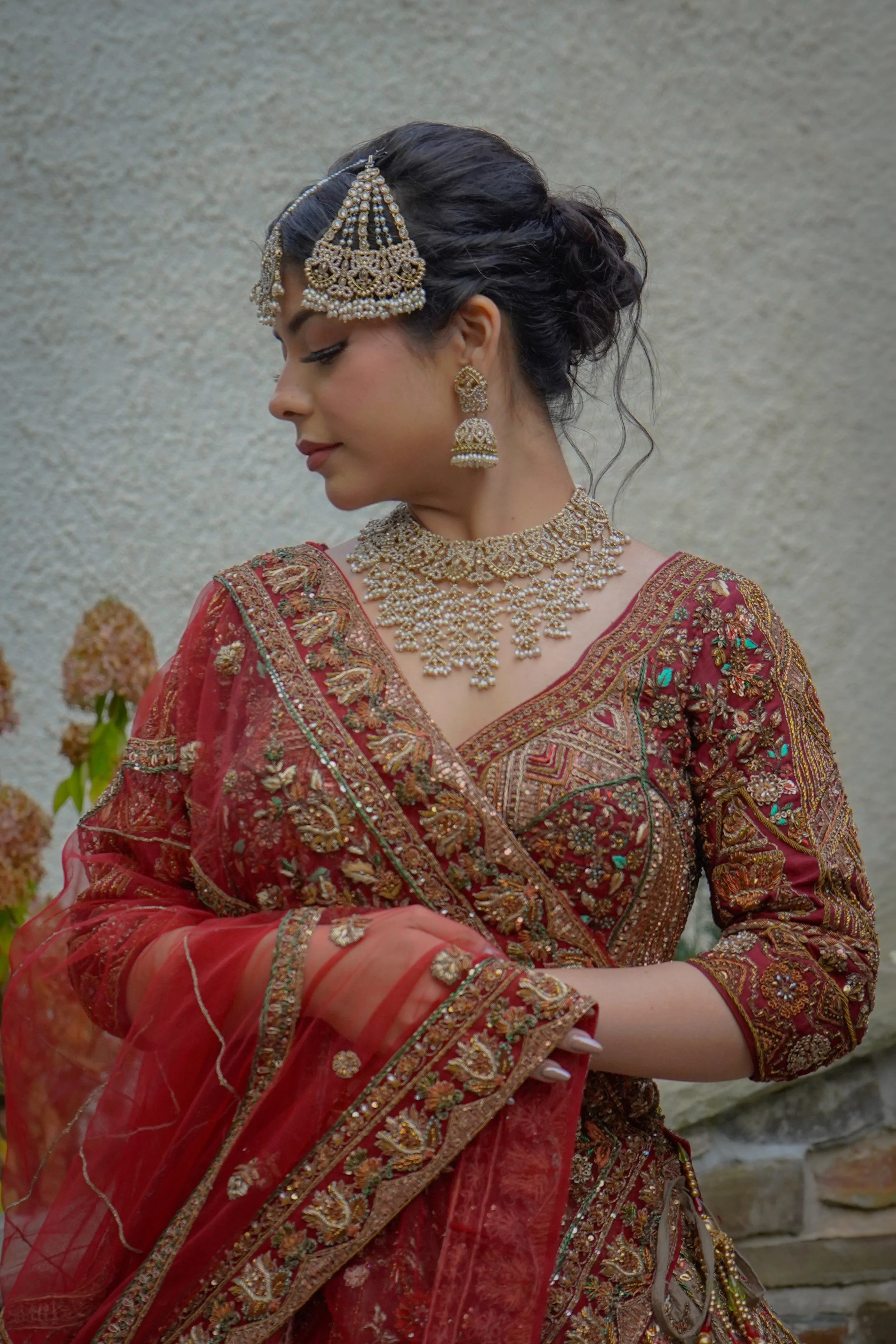A woman dressed in a traditional red and gold embellished outfit, wearing ornate jewelry including a maang tikka on her forehead, large earrings, and multiple layered necklaces, standing outdoors against a neutral background.