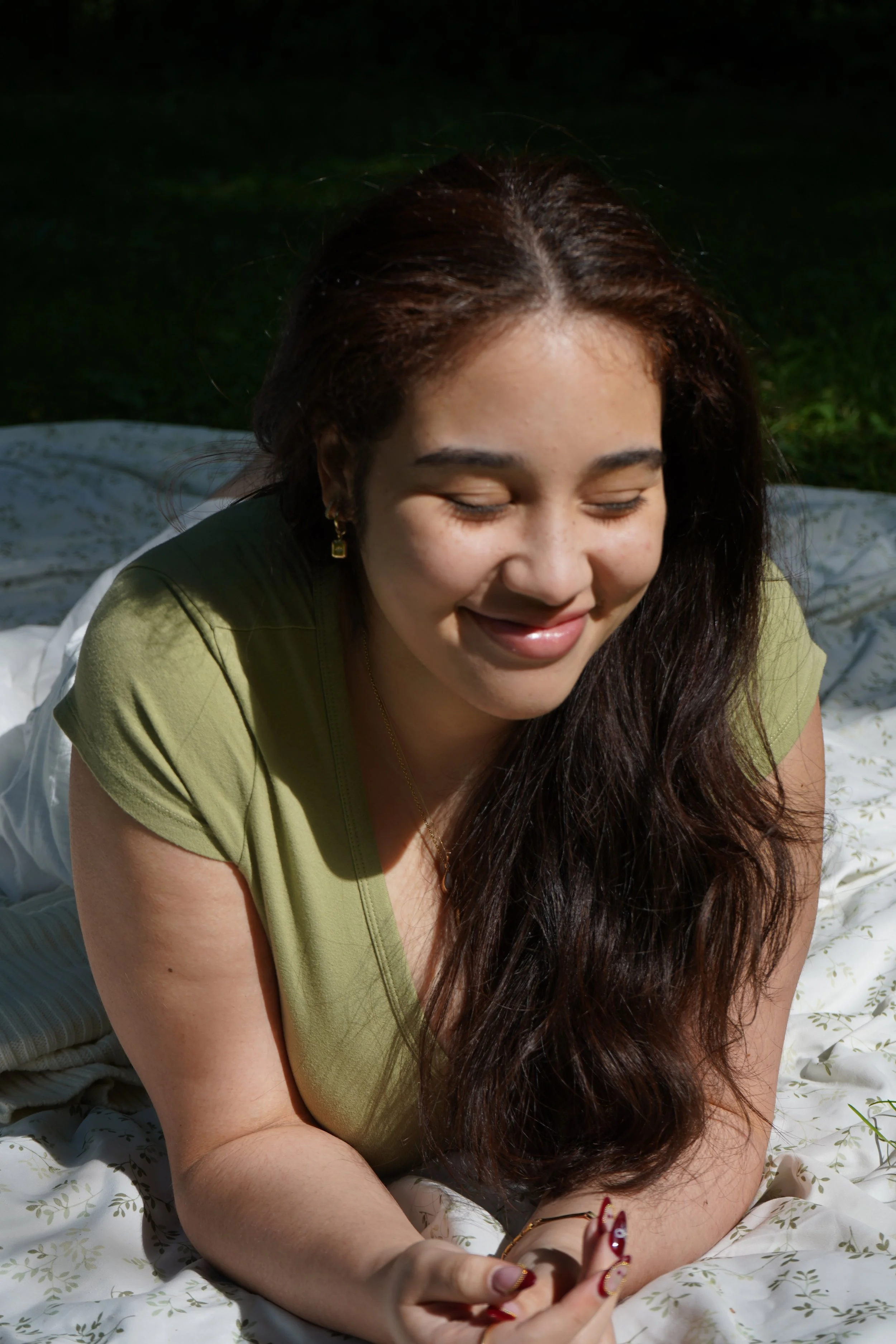 A woman with long dark hair lying on a white floral blanket outdoors, smiling with eyes closed.