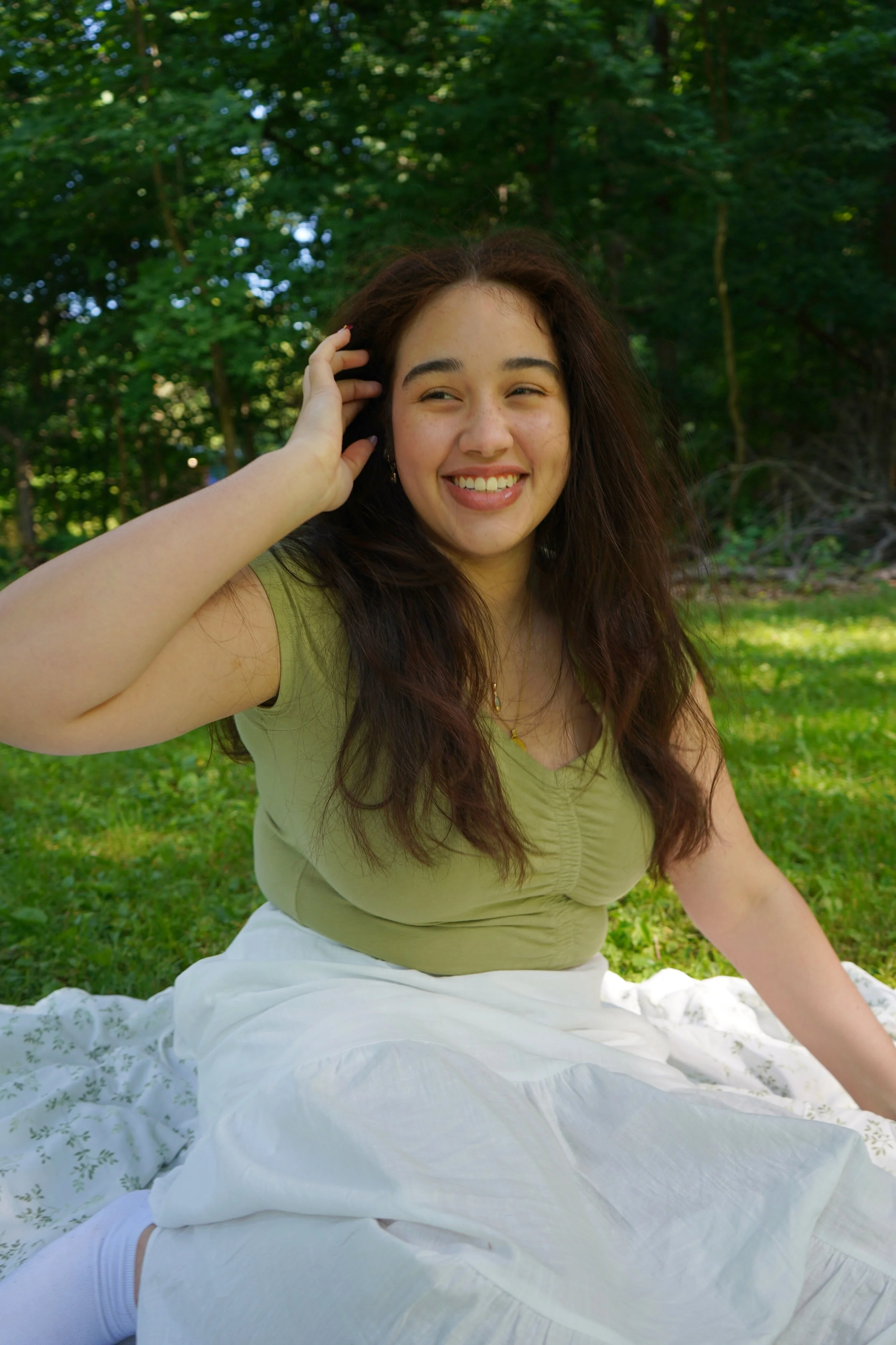 A young woman with long, curly brown hair smiling outdoors, sitting on a white blanket on grass, with trees in the background.