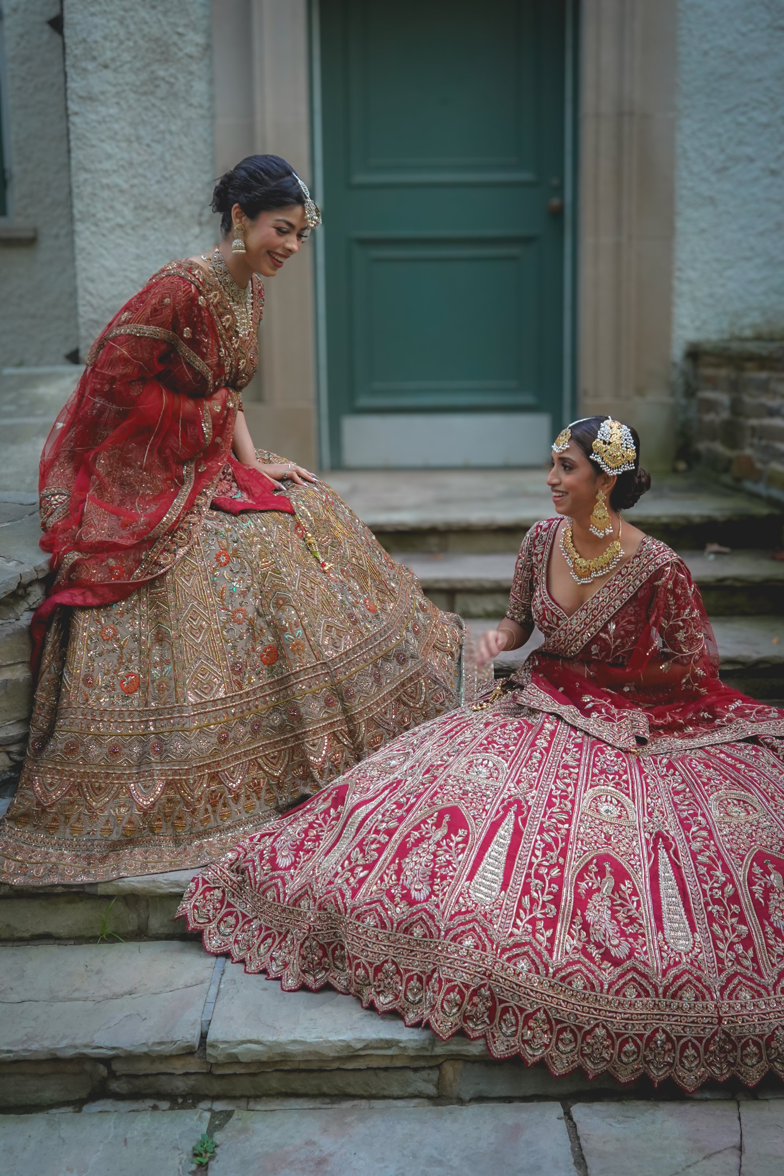 Two women in traditional South Asian bridal attire, dressed in red and gold heavily embroidered gowns, sitting on stone steps outside a building with a green door, sharing a joyful moment.