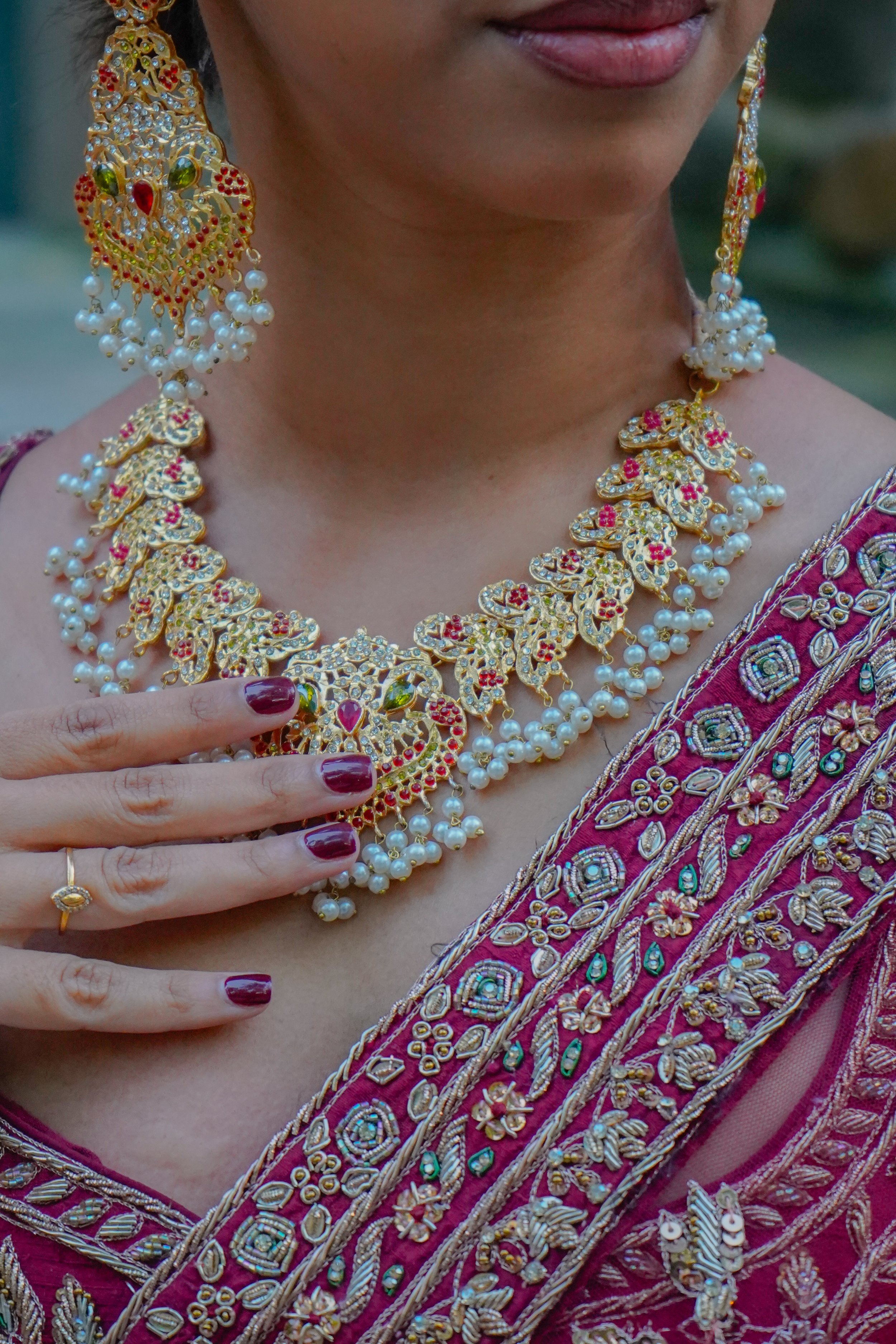 Close-up of a woman wearing ornate gold jewelry with pearls and red and green gemstones, and a richly embroidered pink saree with gold threadwork.