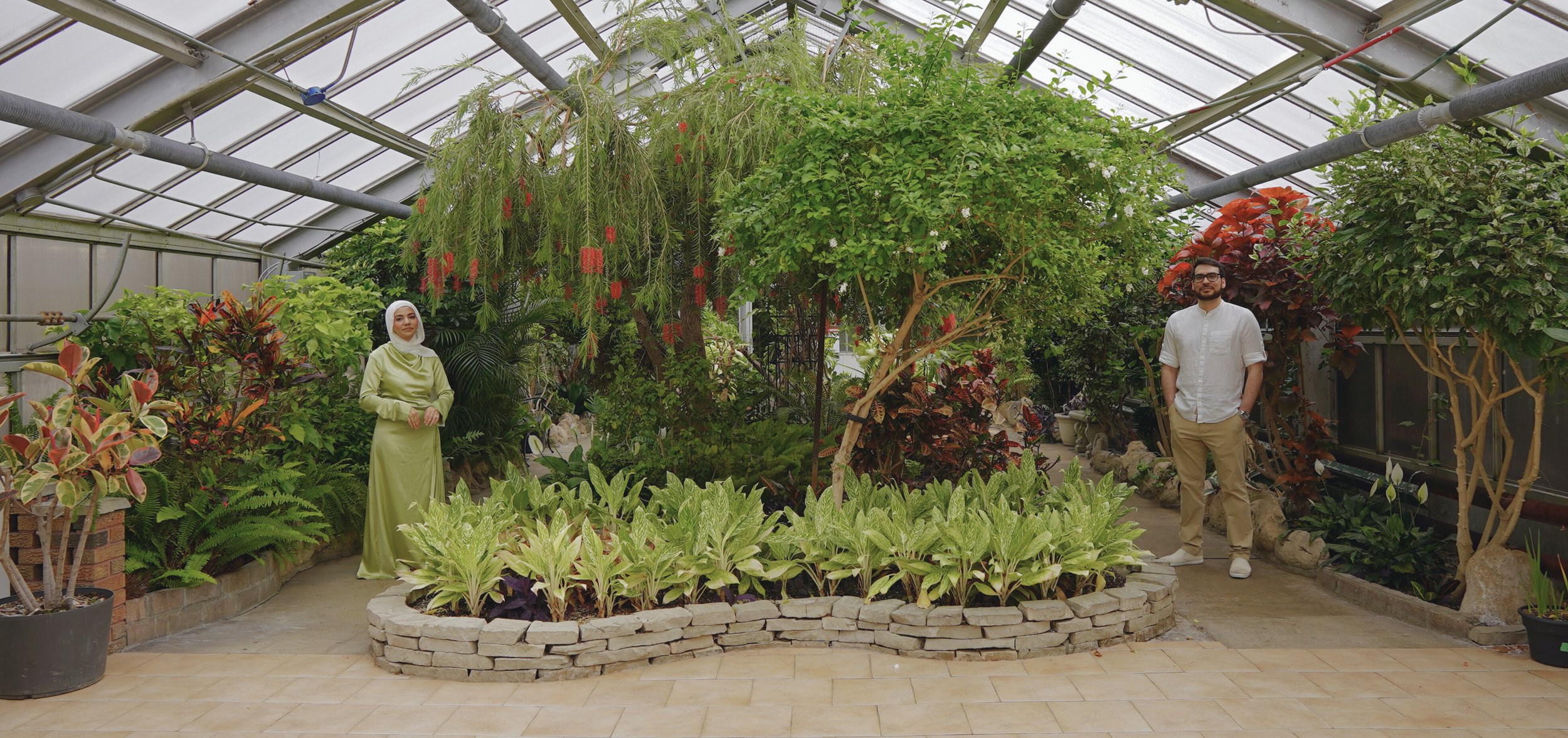 Two people standing inside a greenhouse with lush green plants and trees, one woman wearing a green dress and white headscarf, one man in a white shirt and khaki pants.