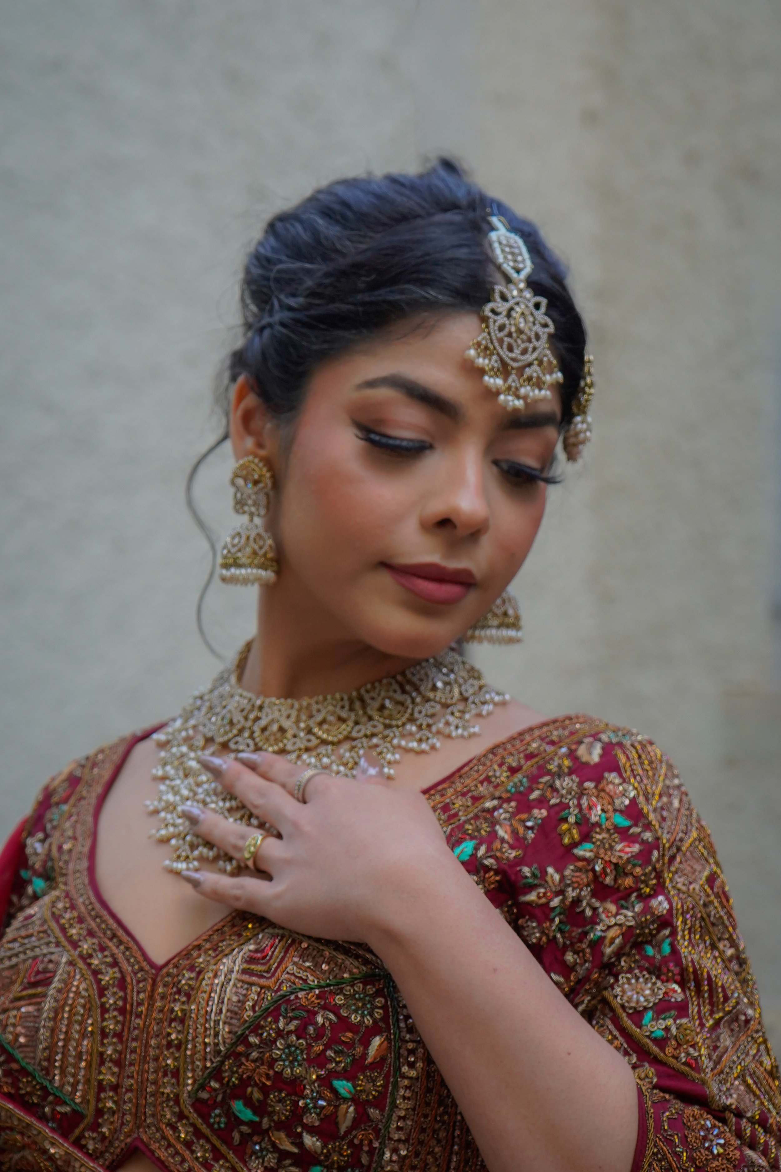 A woman dressed in traditional Indian attire with elaborate gold jewelry, including earrings, a necklace, and a maang tikka, wearing a richly embroidered maroon dress.